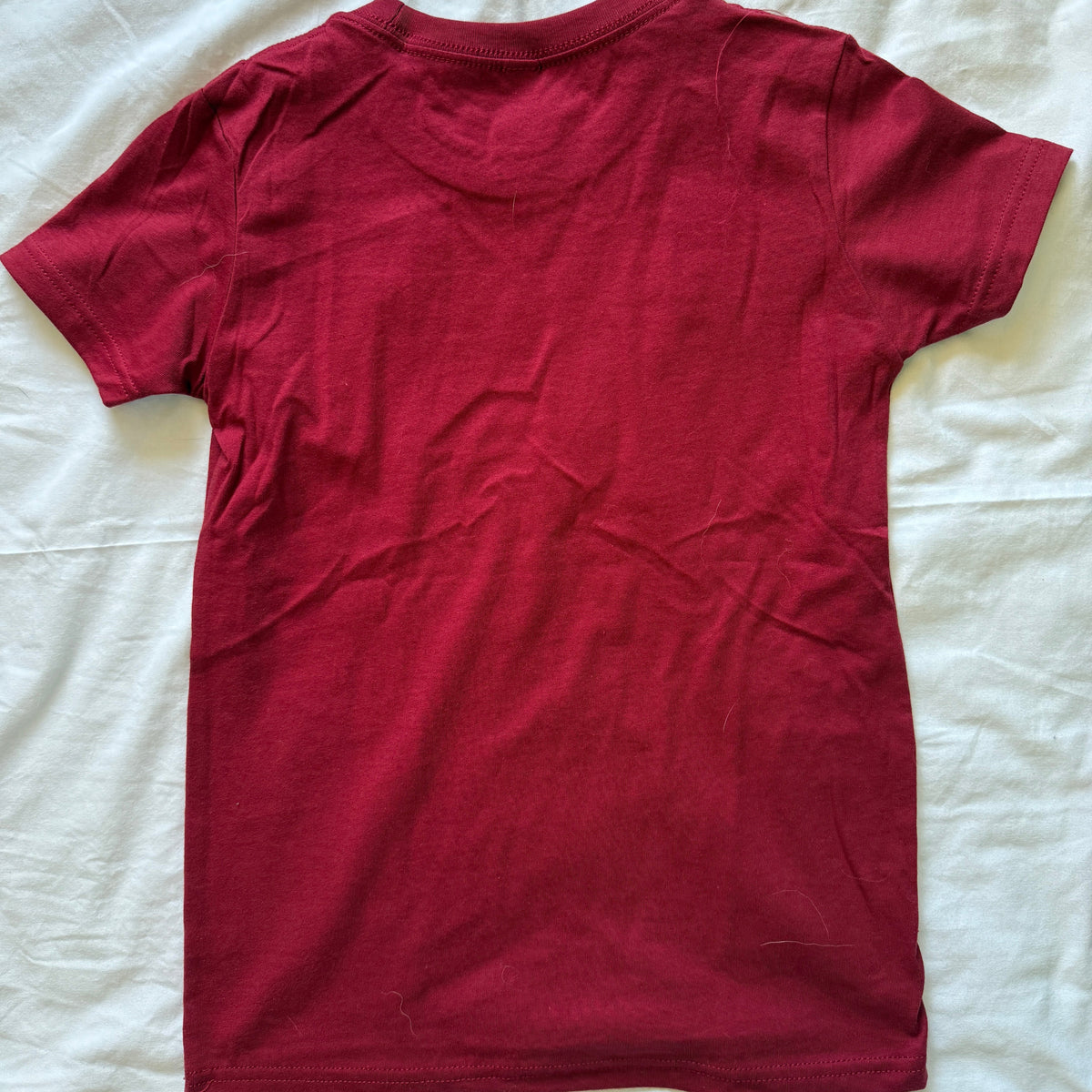 A dark red short-sleeve t-shirt laid flat on a wrinkled white sheet.