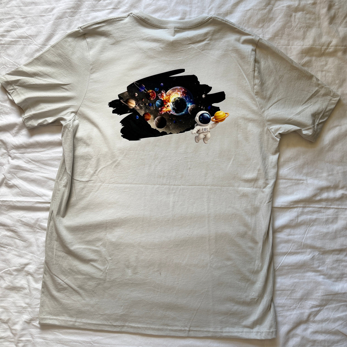 A light beige crewneck t-shirt laid flat on a wrinkled white sheet, featuring a colorful cosmic graphic print on the chest with a small astronaut detail.