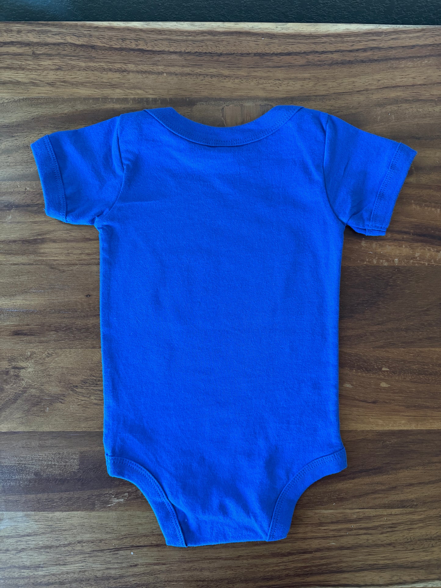 A bright royal blue short-sleeve baby bodysuit laid flat on a dark brown wooden surface.
