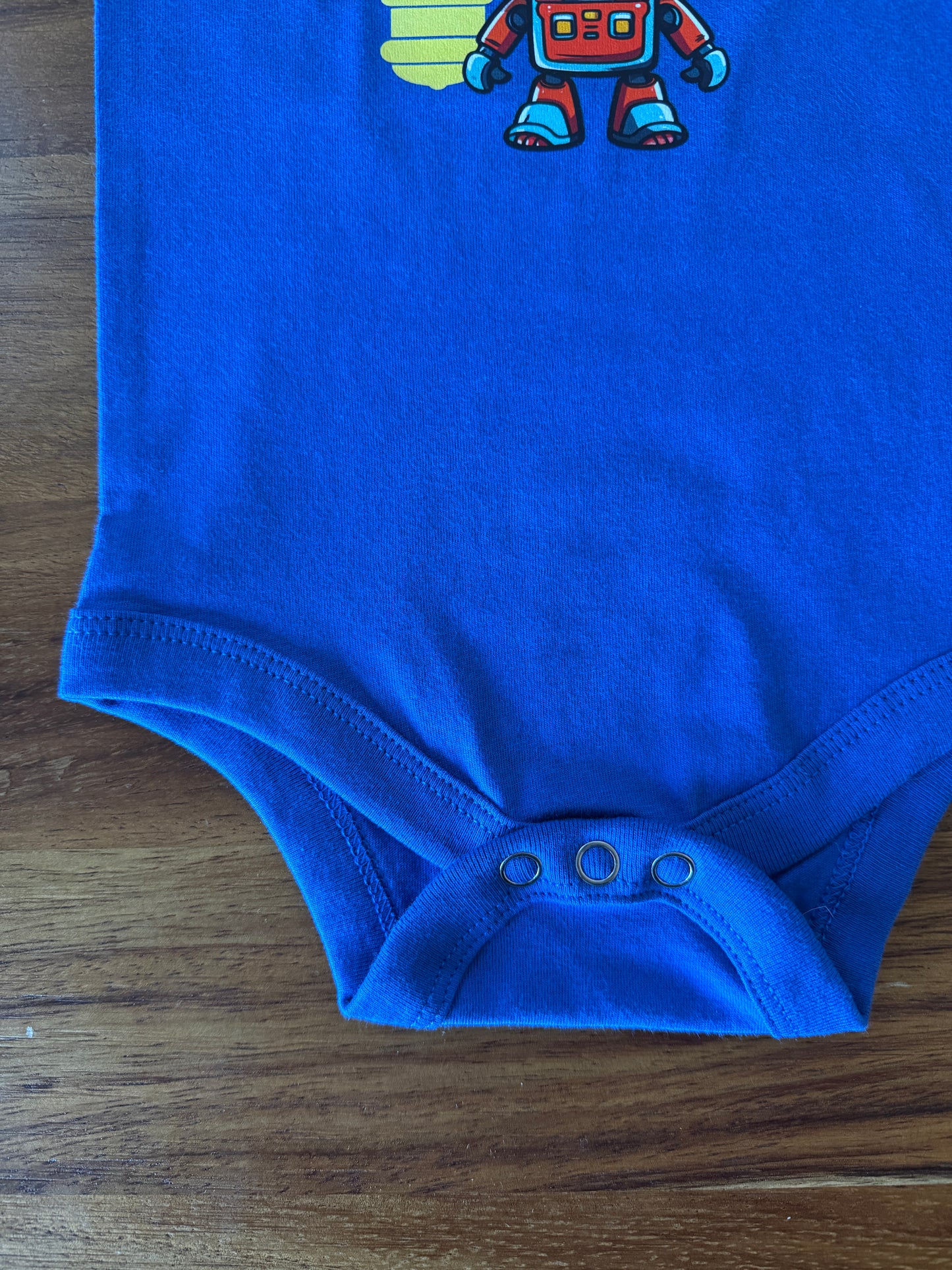 A bright blue baby bodysuit laid flat on a wooden surface, featuring a small colorful robot graphic near the neckline and three snap closures at the bottom.