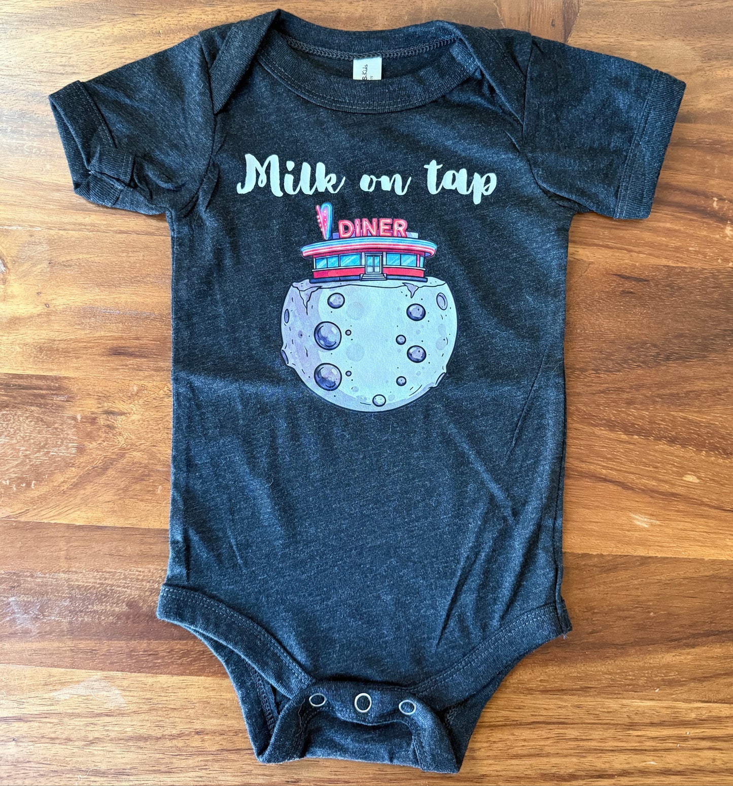 A dark heather gray baby bodysuit laid flat on a wooden surface, featuring a graphic of a retro diner on a moon with the text 'Milk on tap'.