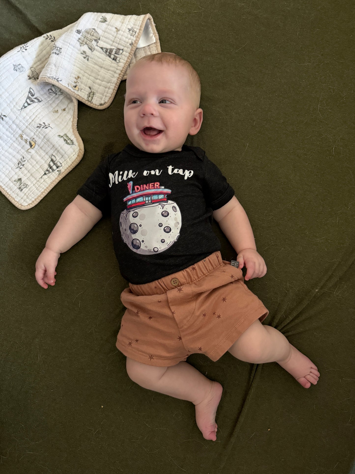 A baby lying on an olive green blanket, wearing a black short-sleeve t-shirt with a moon graphic and the text 'Milk on tap Diner' paired with tan shorts.