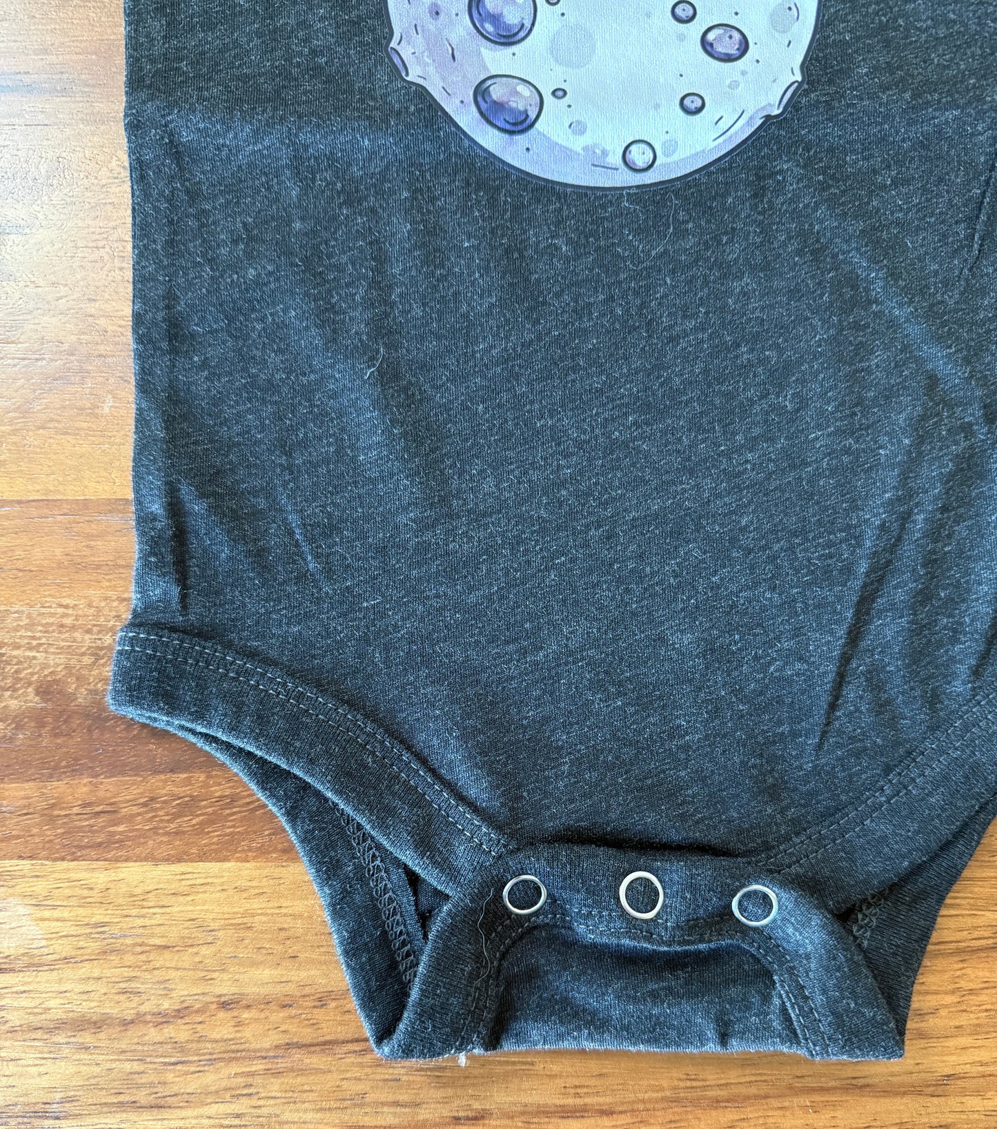 A dark charcoal gray baby bodysuit laid flat on a wooden surface, featuring a partial graphic of a moon with purple and blue craters at the top and three silver snap closures at the bottom.