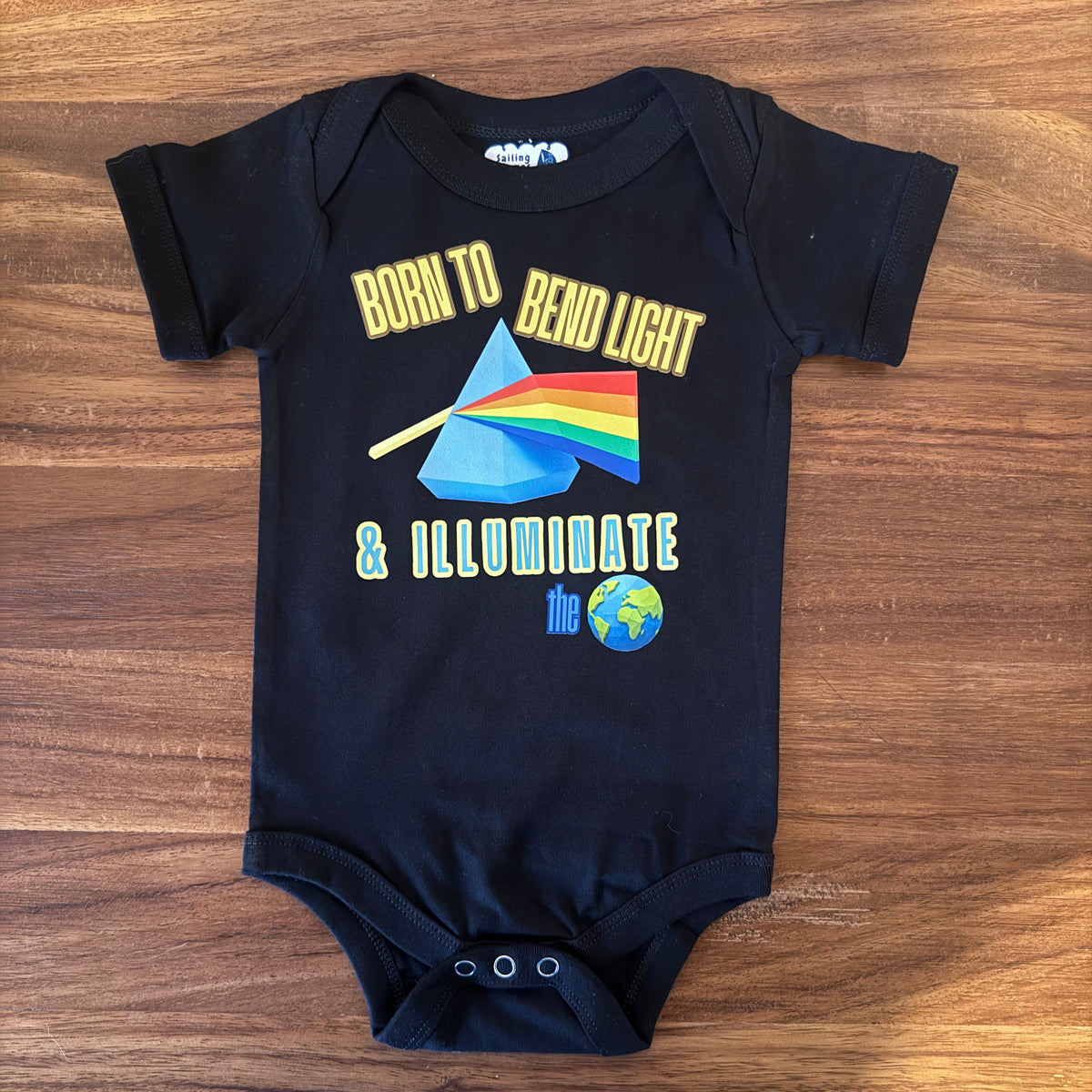 A black short-sleeve baby bodysuit laid flat on a wooden surface, featuring a colorful graphic print with the text 'BORN TO BEND LIGHT & ILLUMINATE the globe' and a prism design.