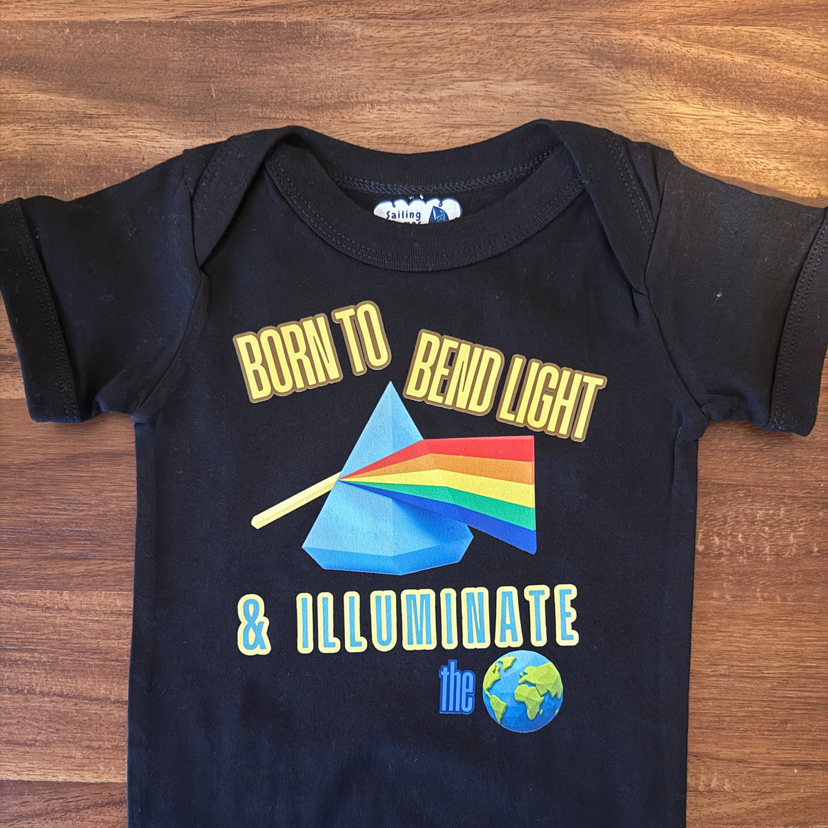 A black baby bodysuit laid flat on a wooden surface, featuring a graphic print with the text 'BORN TO BEND LIGHT & ILLUMINATE the globe' and a prism illustration in rainbow colors.