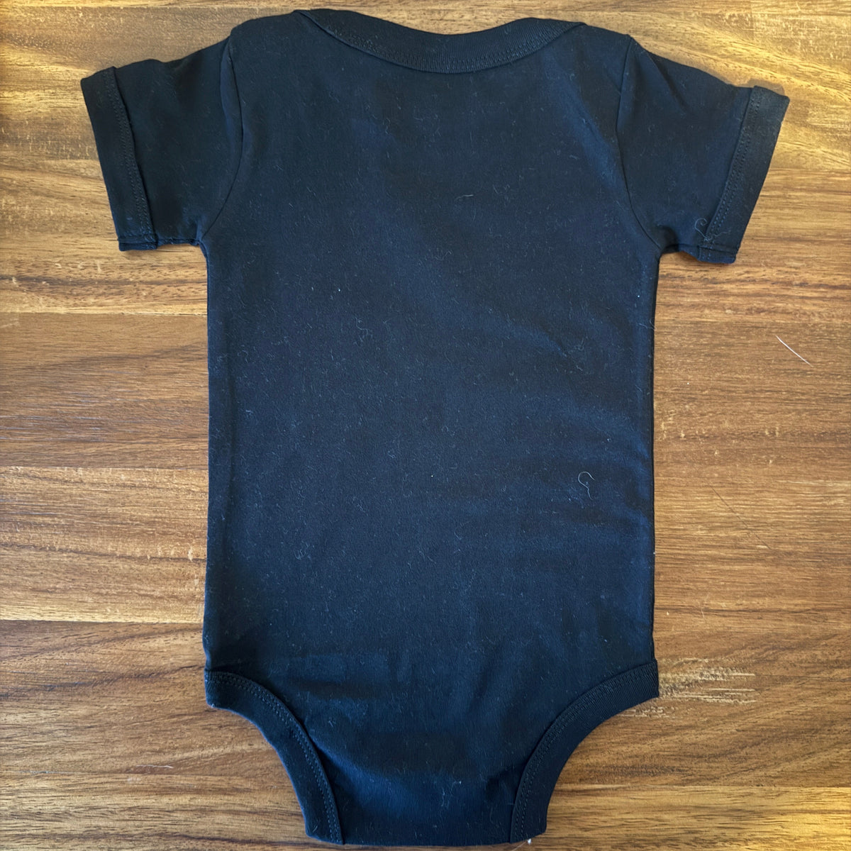 A plain black short-sleeve baby bodysuit laid flat on a wooden table.