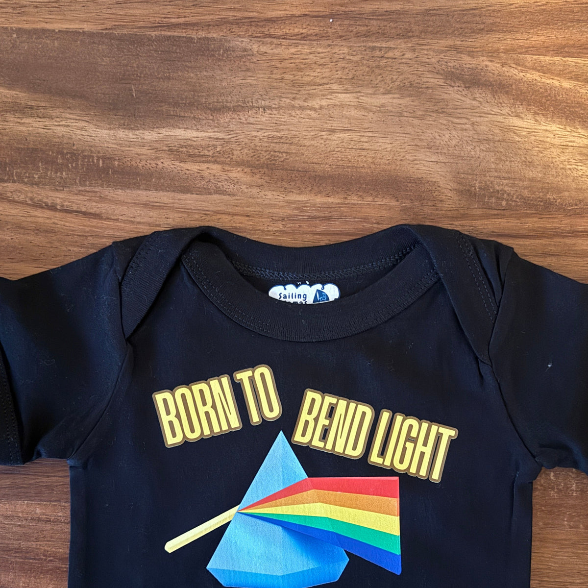 A black short-sleeve baby bodysuit laid flat on a wooden surface, featuring a colorful graphic print with the text 'BORN TO BEND LIGHT & ILLUMINATE the globe' and a prism made from a rainbow flag.