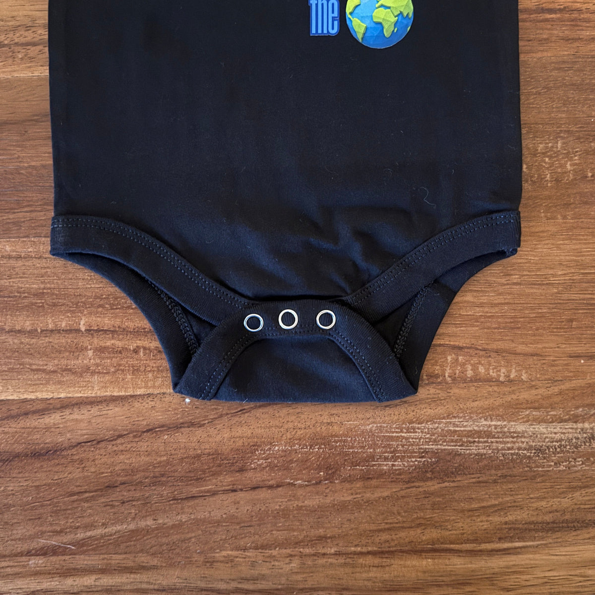 A black baby bodysuit laid flat on a wooden surface, featuring a Pink Floyd 'The Dark Side of the Moon' themed graphic with the text 'BORN TO BE DARK LIGHT & ILLUMINATE' and a prism illustration.