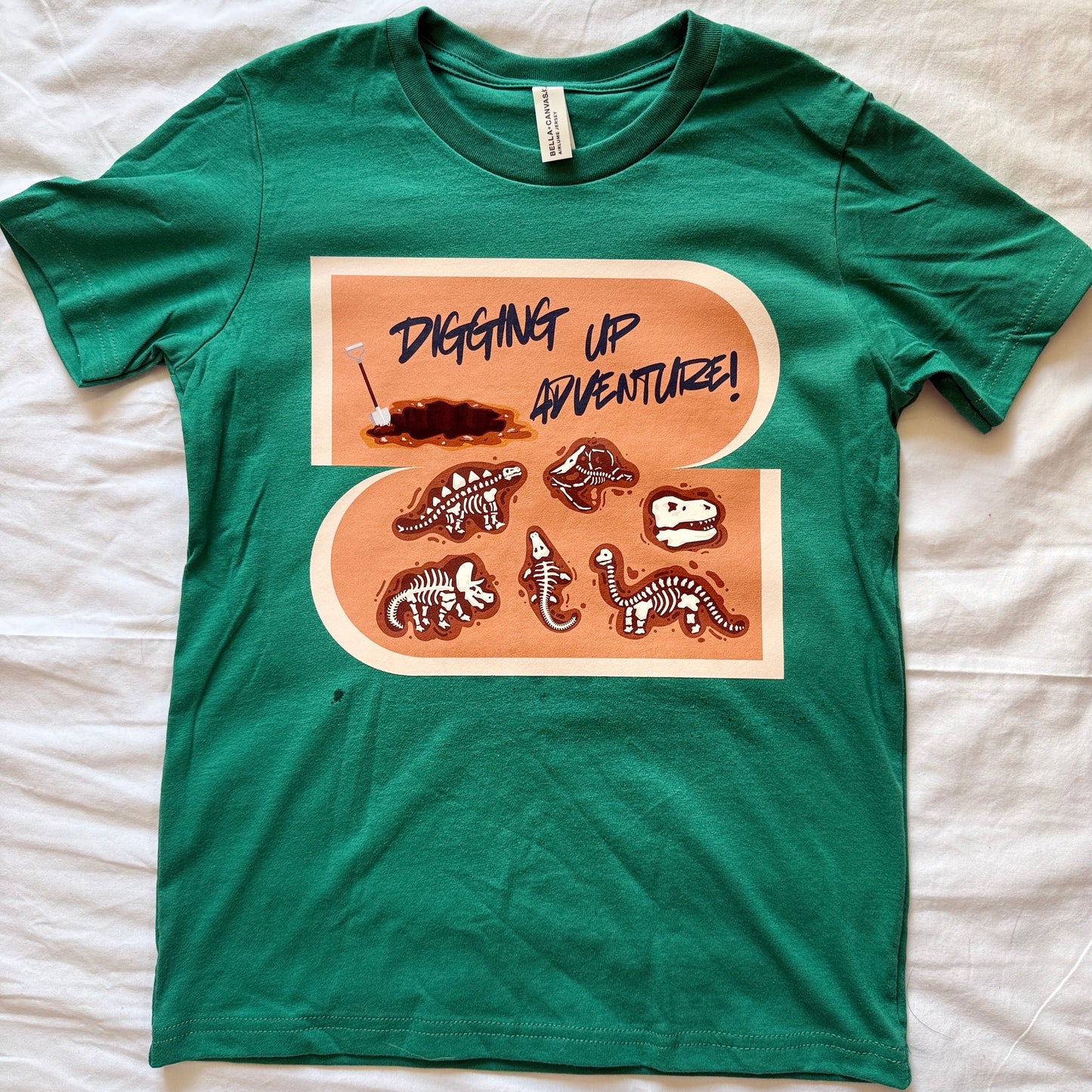 A green short-sleeve t-shirt laid flat on a white fabric background, featuring a large peach-colored graphic with dinosaur bones and the text 'DIGGING UP ADVENTURE!'.