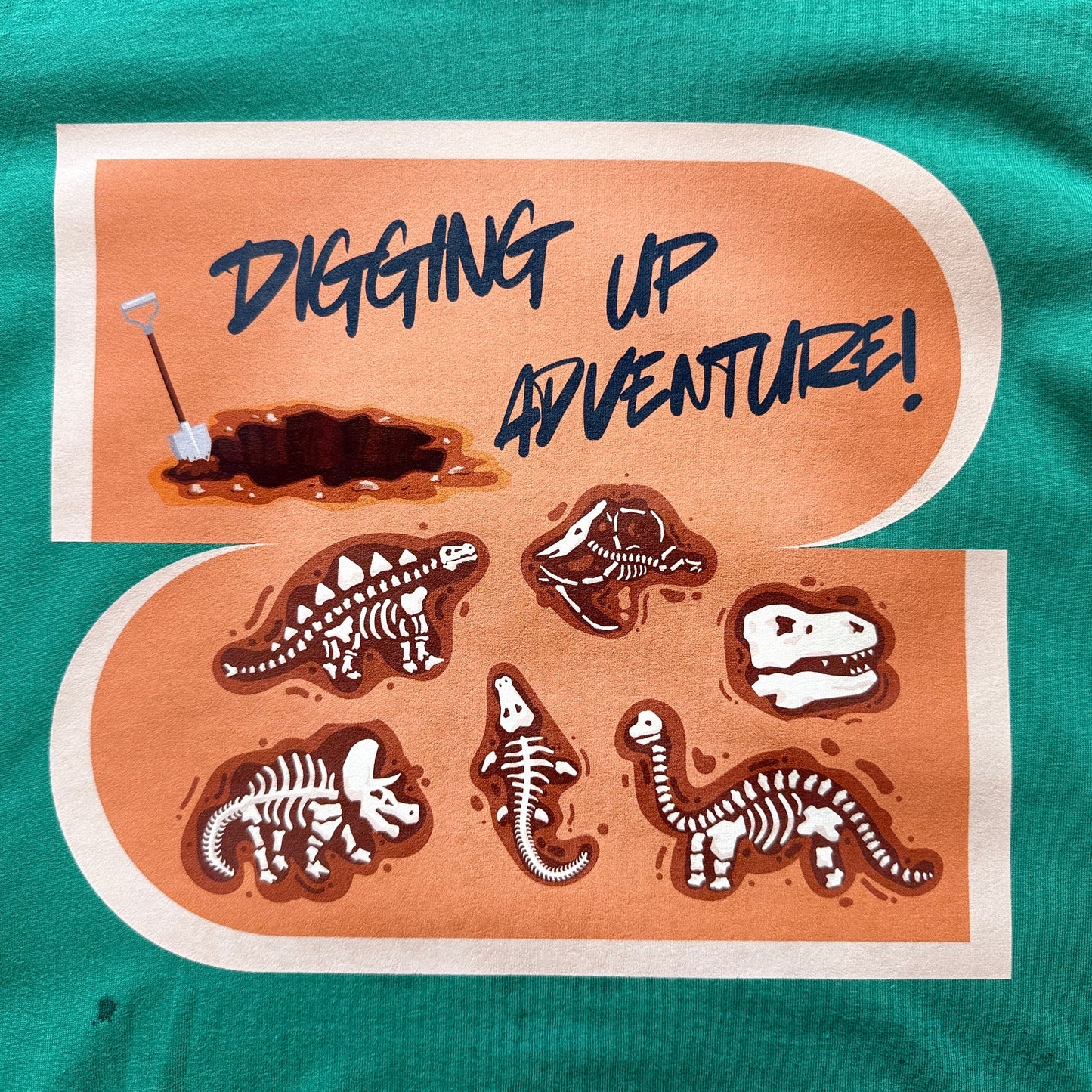 A close-up of a green t-shirt featuring a large graphic print with the text 'DIGGING UP ADVENTURE!' and illustrations of dinosaur skeletons and a hole being dug.