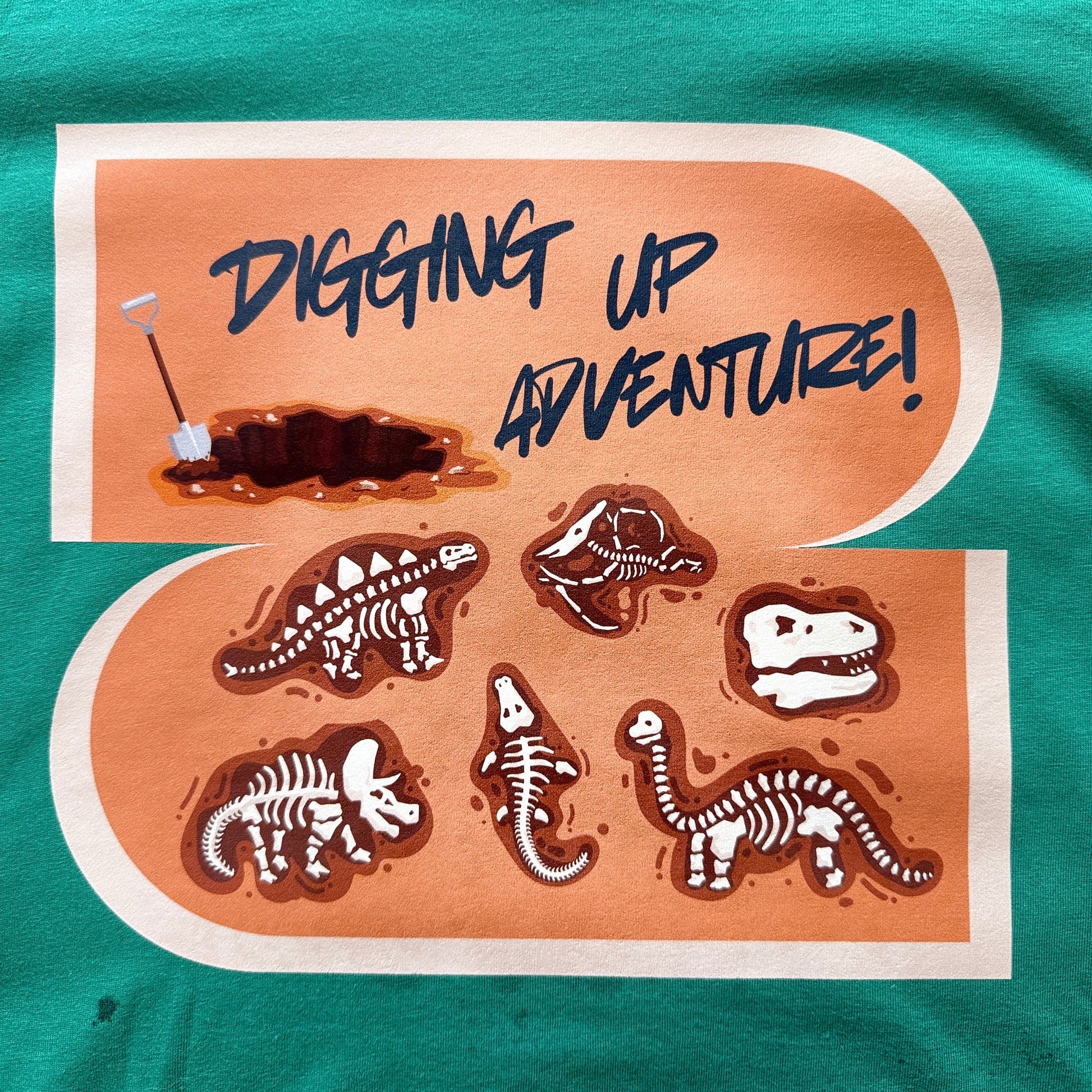 A close-up of a green t-shirt featuring a large graphic print with the text 'DIGGING UP ADVENTURE!' and illustrations of dinosaur skeletons and a hole being dug.