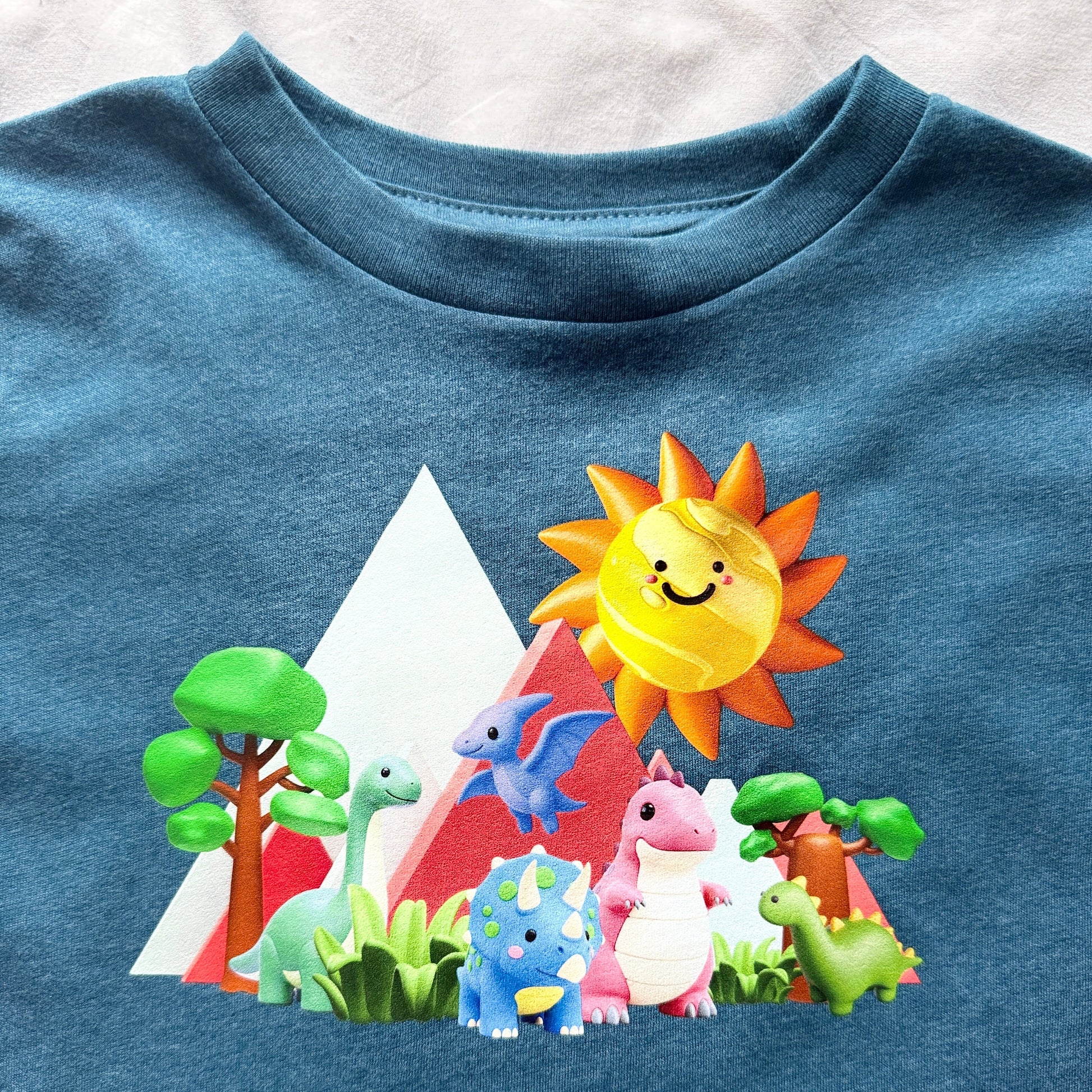 A close-up of a blue heathered t-shirt featuring a colorful cartoon graphic of dinosaurs, a smiling sun, and mountains.