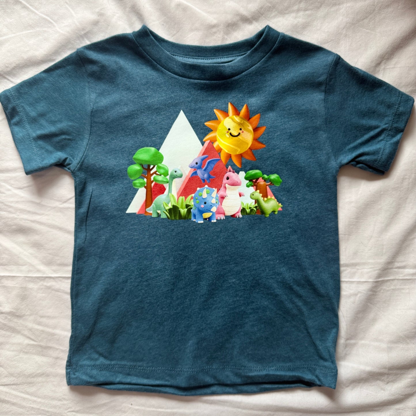 A toddler's heathered teal blue short-sleeve t-shirt with a colorful cartoon graphic of a sun, mountains, and cute creatures, laid flat on a wrinkled off-white sheet.