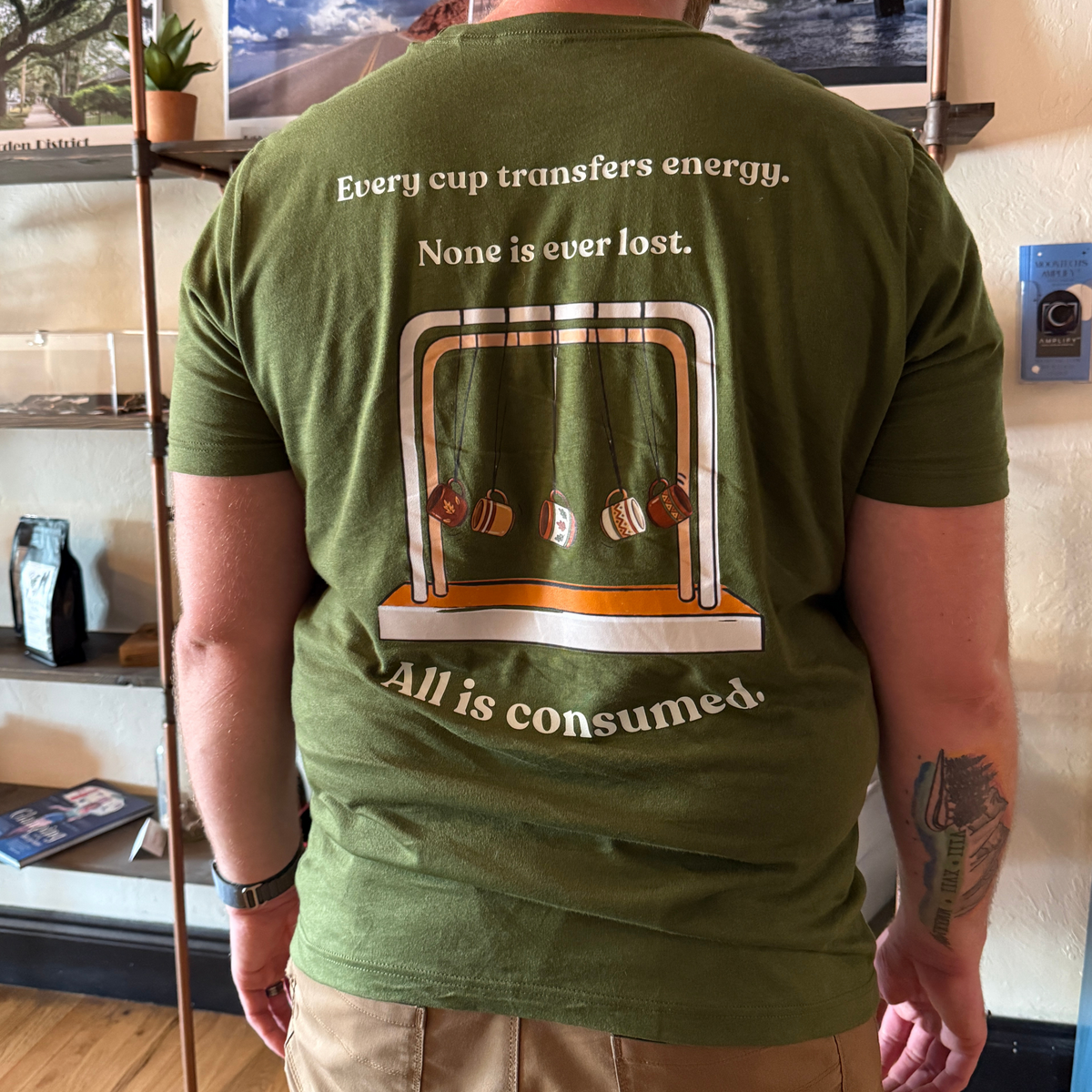 A man viewed from behind wearing an olive green t-shirt with a back graphic that reads 'Every cup transfers energy. None is ever lost.