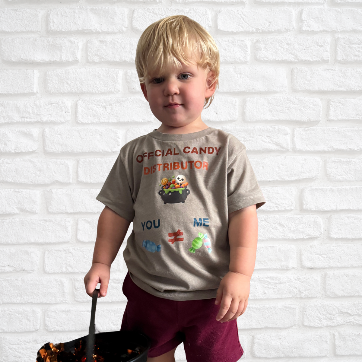 A young blonde boy wearing a beige graphic t-shirt that reads 'OFFICIAL CANDY DISTRIBUTOR' and holding a black bucket, standing in front of a white brick wall.