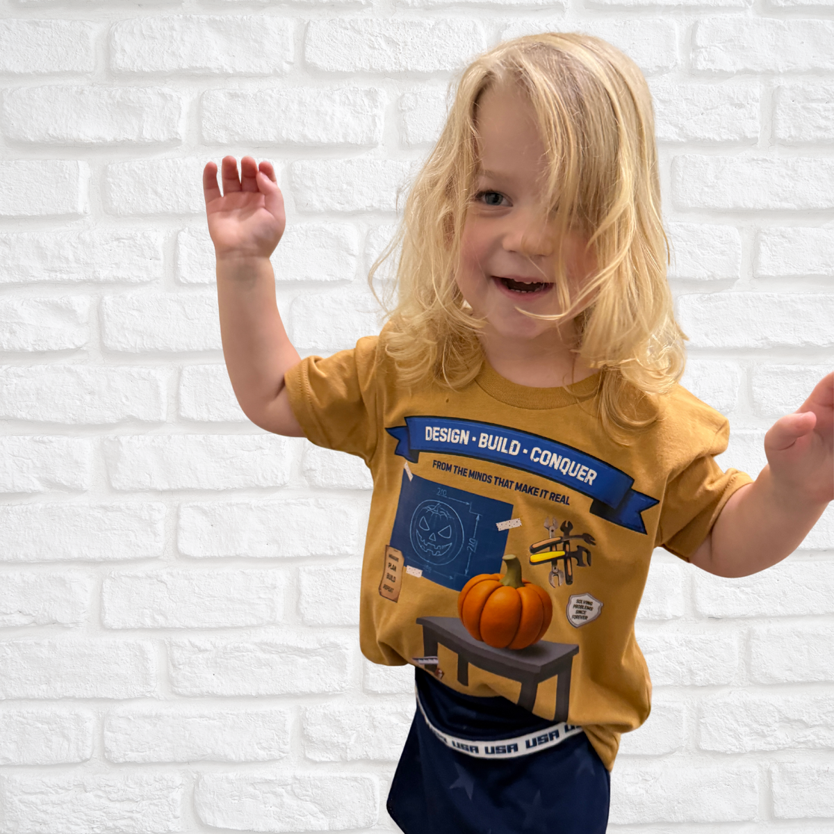 A young child with blonde hair wearing a mustard yellow graphic t-shirt and navy blue pants, standing against a white brick wall and waving.