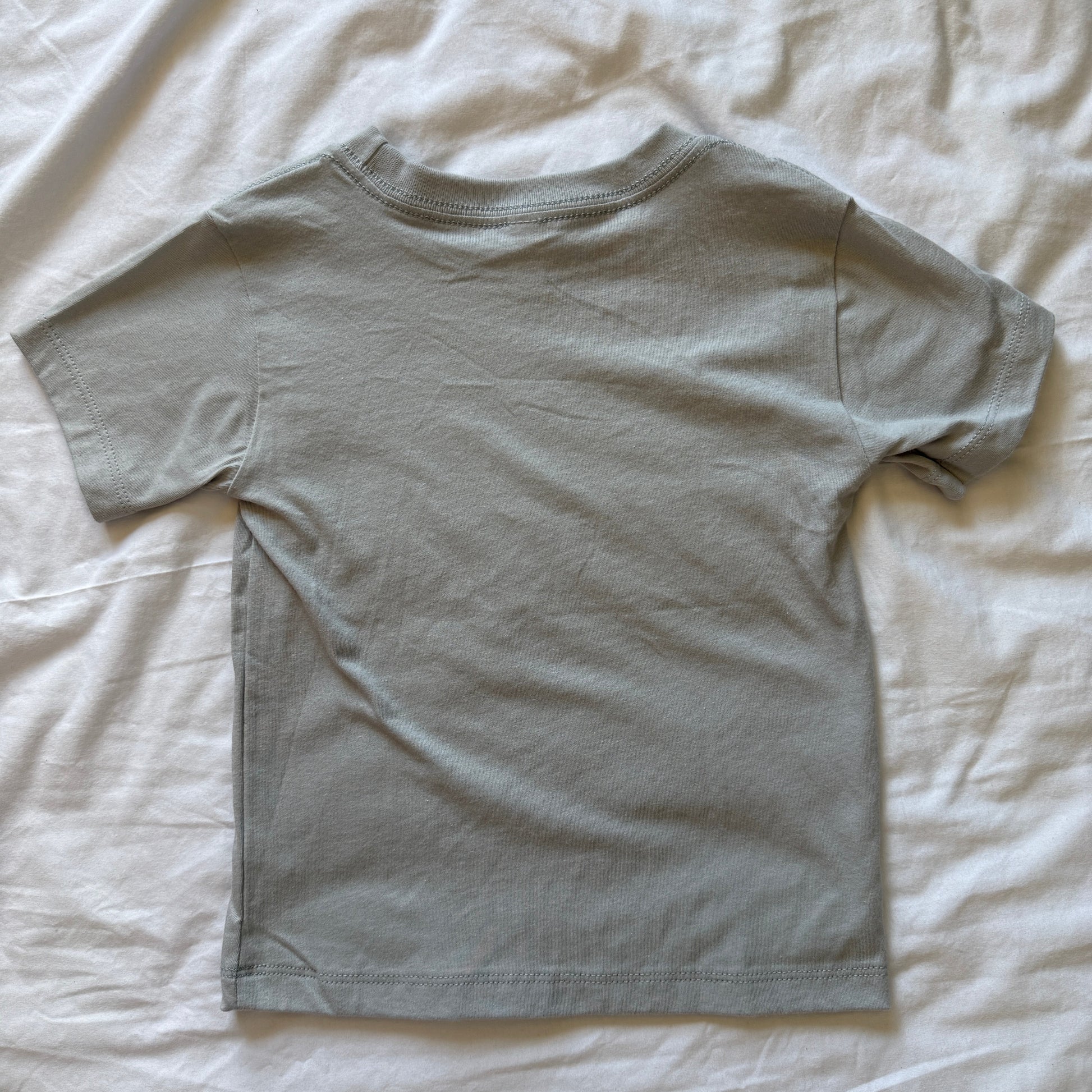 A light gray short-sleeve t-shirt laid flat on a wrinkled white sheet.