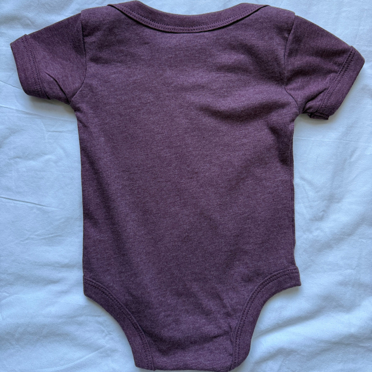 A short-sleeve baby bodysuit in heathered plum purple, laid flat on a wrinkled white sheet.