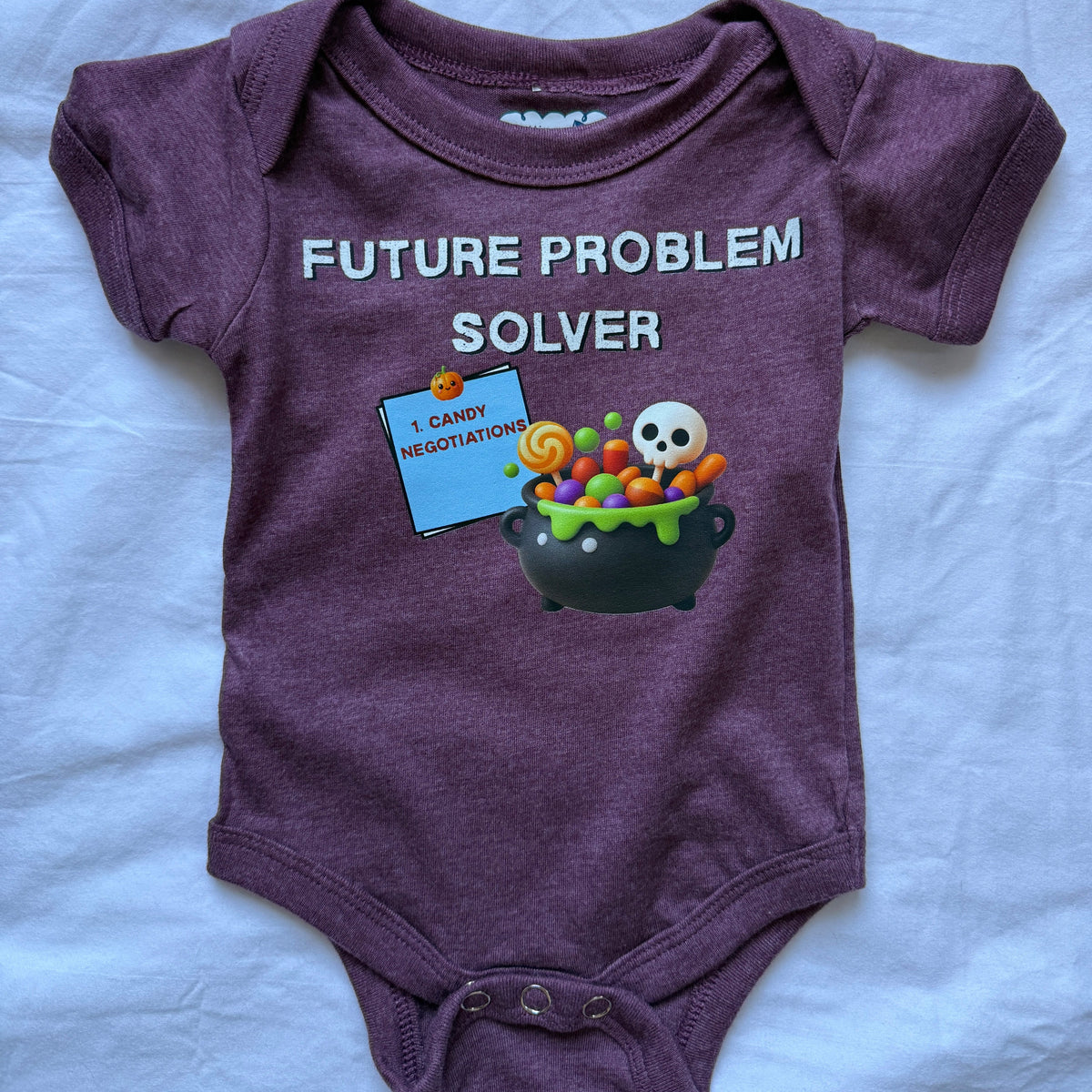 A heathered purple short-sleeve baby bodysuit laid flat on a white fabric background. The bodysuit features a graphic print with the text 'FUTURE PROBLEM SOLVER' in white, a blue sign reading 'I CARRY NEGOTIATION', and a black cauldron filled with colorful candies, a green slime, and a white skull.