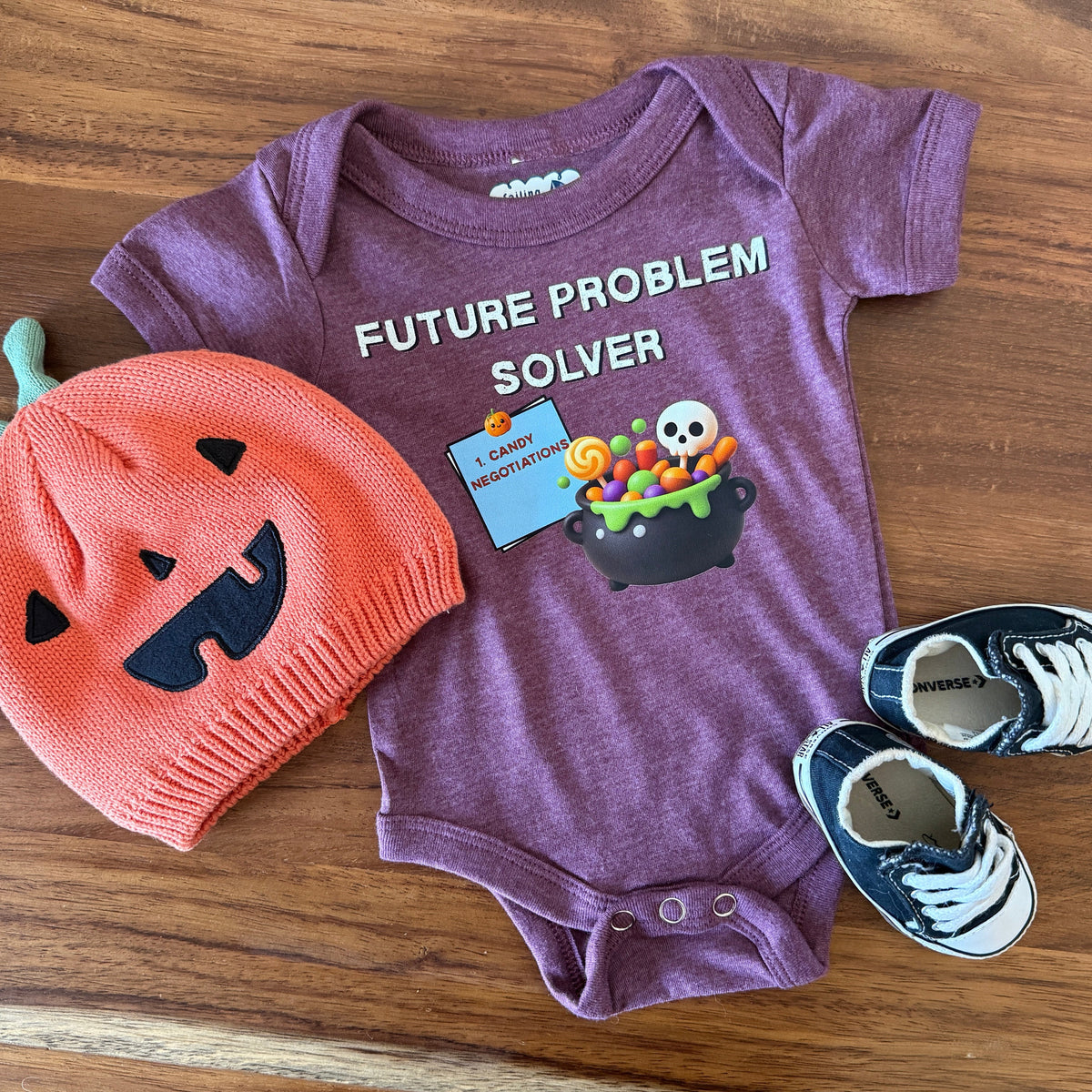A baby clothing set laid flat on a wooden floor, including a heathered purple long-sleeve bodysuit with a Halloween graphic and the text 'FUTURE PROBLEM SOLVER,' a coral-orange knit pumpkin hat, and navy sneakers with white laces.