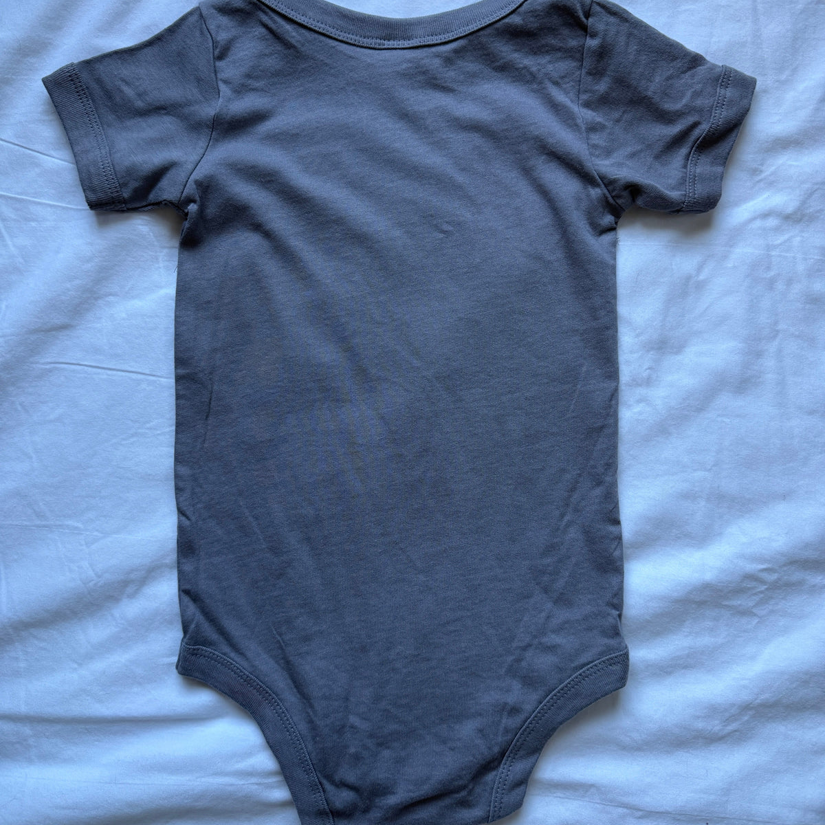 A dark gray short-sleeve baby bodysuit laid flat on a light blue-gray wrinkled fabric background.