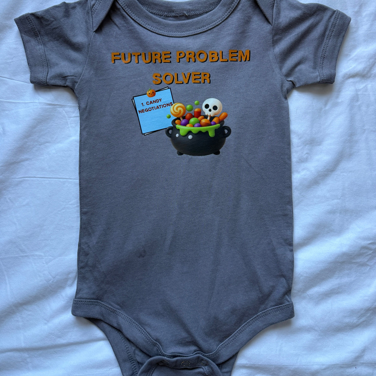 A dark gray short-sleeve baby bodysuit laid flat on a light blue fabric background. The bodysuit features a graphic print with the text 'FUTURE PROBLEM SOLVER' in orange, above a cartoon cauldron filled with candy and a skull.