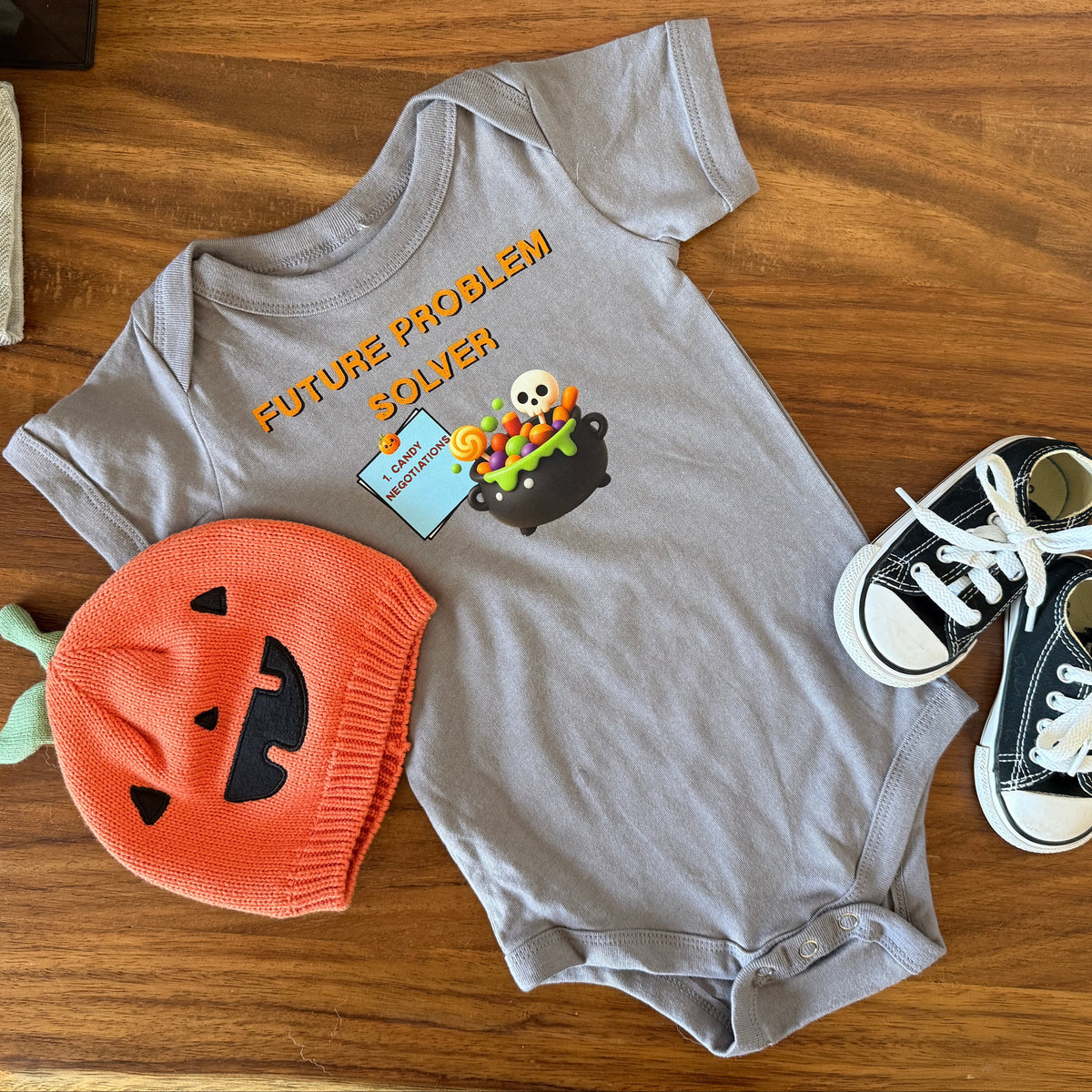A baby clothing flat lay on a wooden surface featuring a heather gray short-sleeve bodysuit with a 'FUTURE PROBLEM BOYZER' Halloween graphic, an orange knit pumpkin hat, and black and white sneakers.