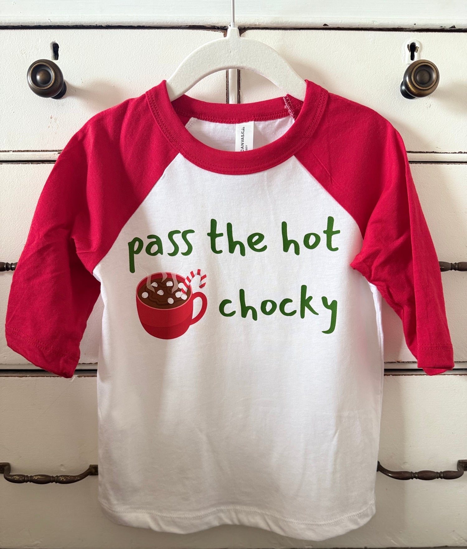 Children's shirt with red sleeves and white body, featuring text and a graphic of hot chocolate on a wooden surface.