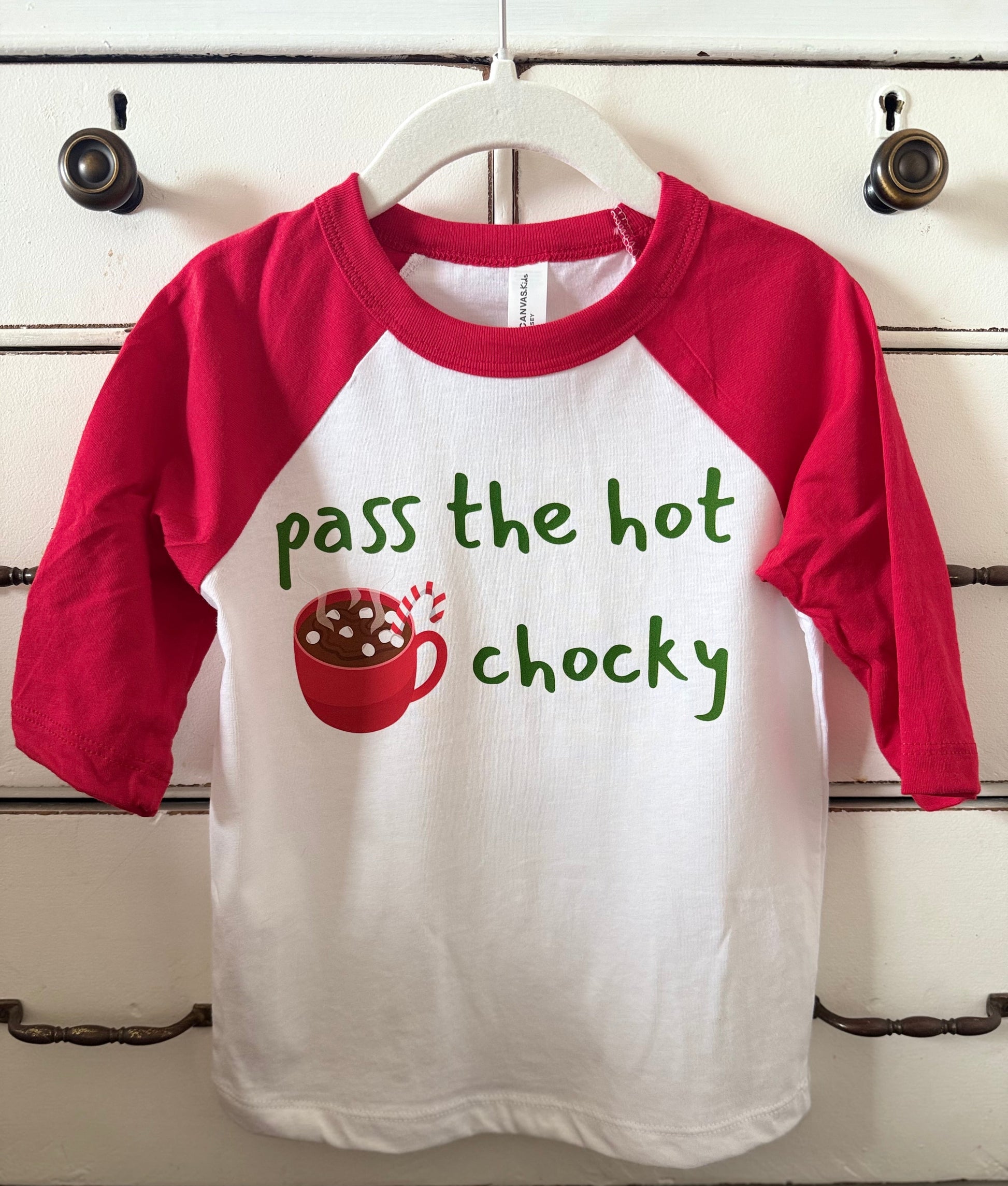 Children's shirt with red sleeves and white body, featuring text and a graphic of hot chocolate on a wooden surface.