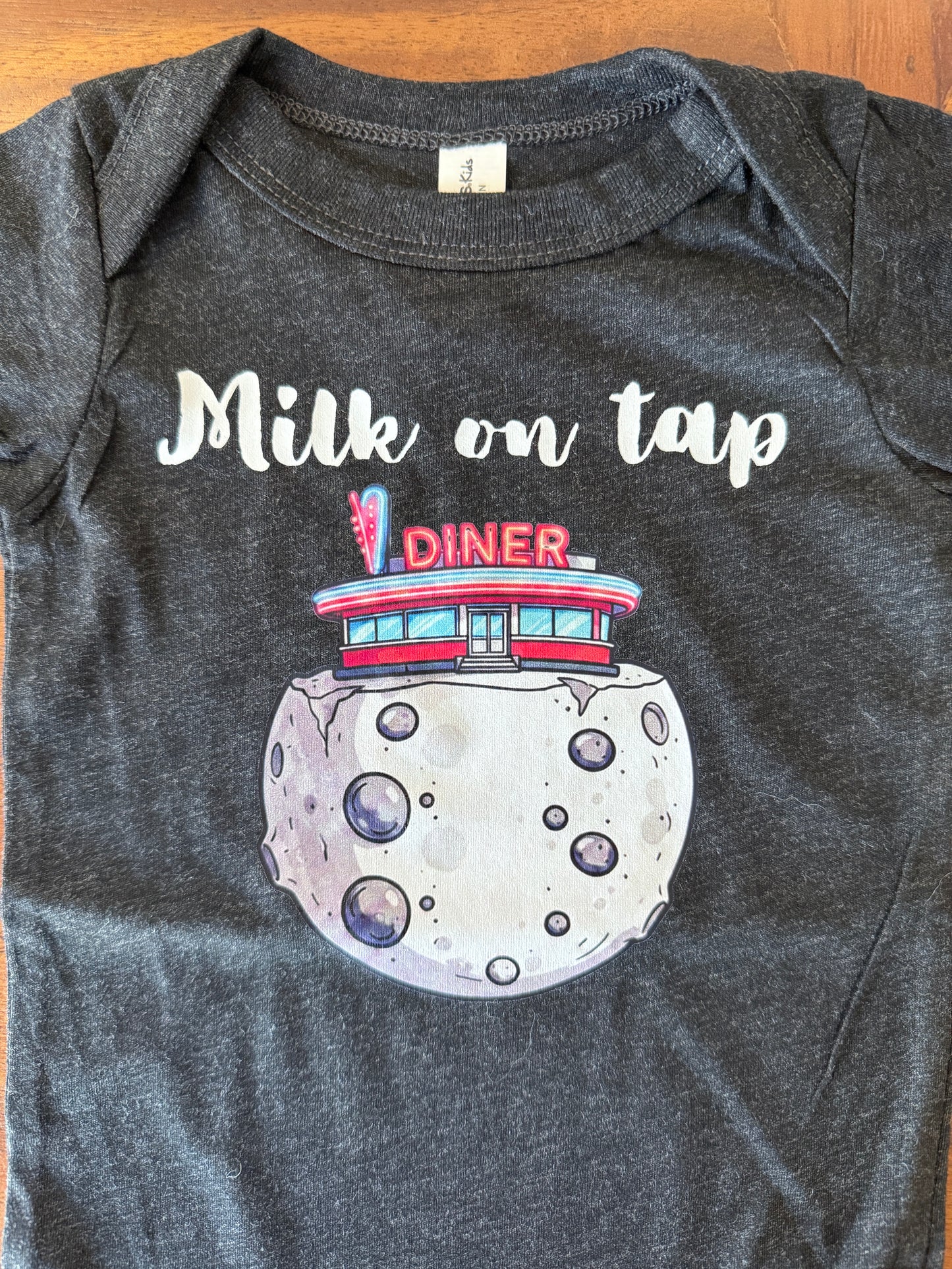 A dark heather gray baby bodysuit laid flat on a wooden surface, featuring a graphic print of a moon with a retro diner on top and the text 'Milk on tap' in white script.