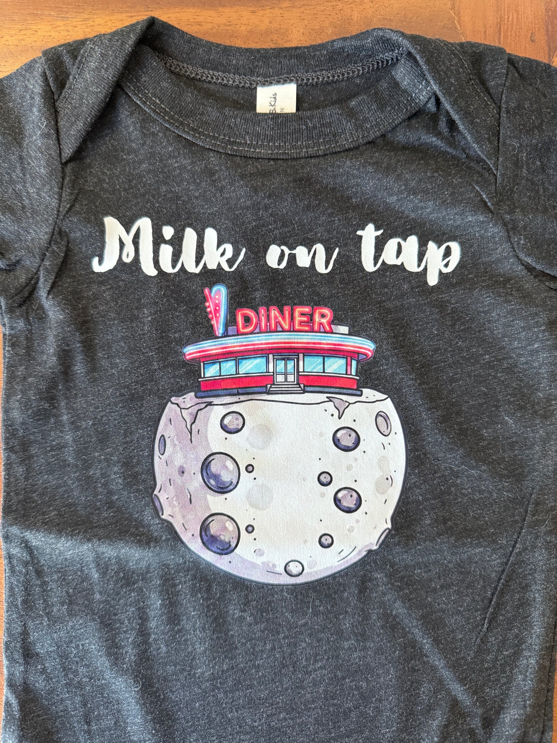 A dark heather gray baby bodysuit laid flat on a wooden surface, featuring a graphic print of a moon with a retro diner on top and the text 'Milk on tap' in white script.