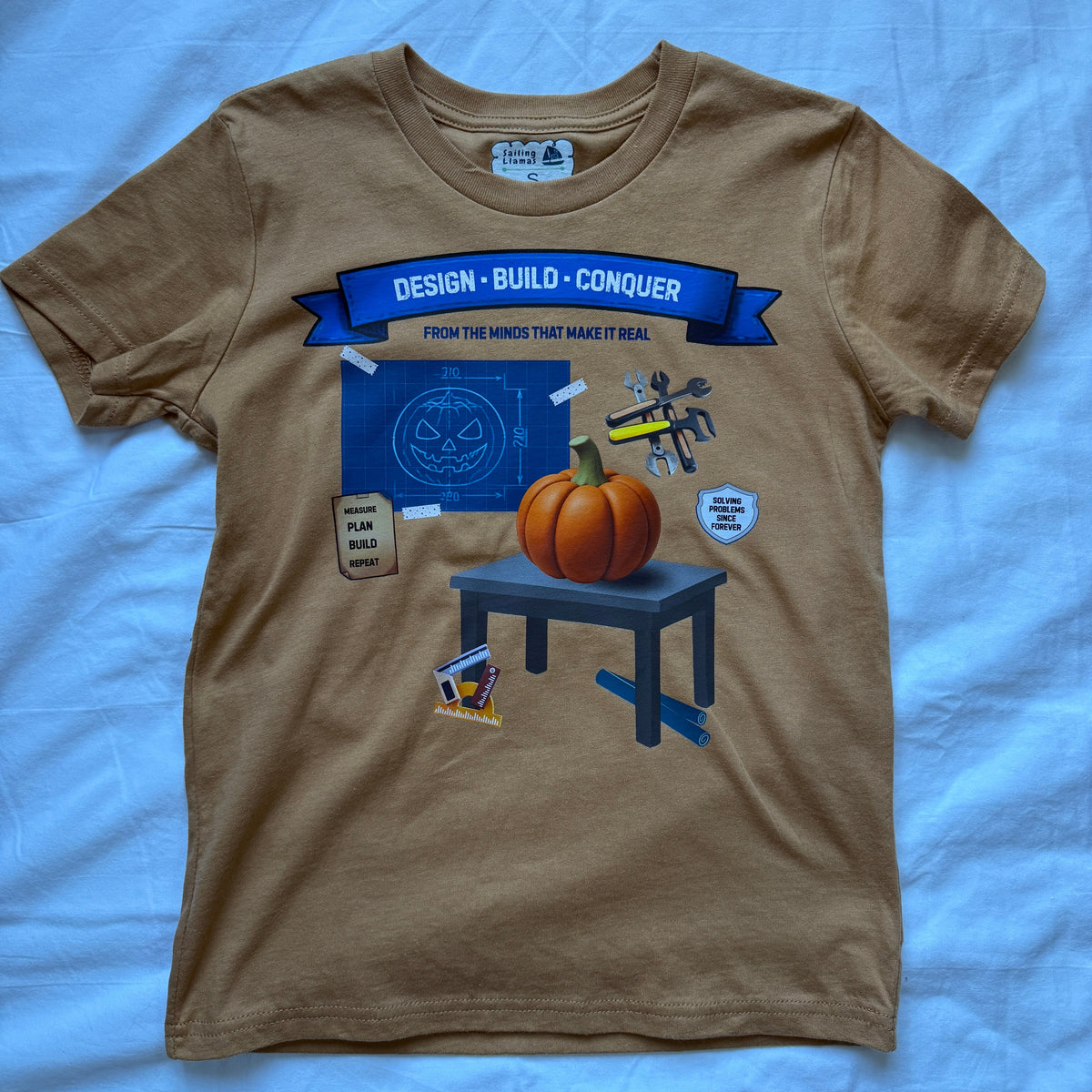 A brown short-sleeve graphic t-shirt laid flat on a light blue fabric background. The graphic features a blue banner reading 'DESIGN • BUILD • CONQUER' and an illustration of a pumpkin on a table with a circuit board and a small robot.