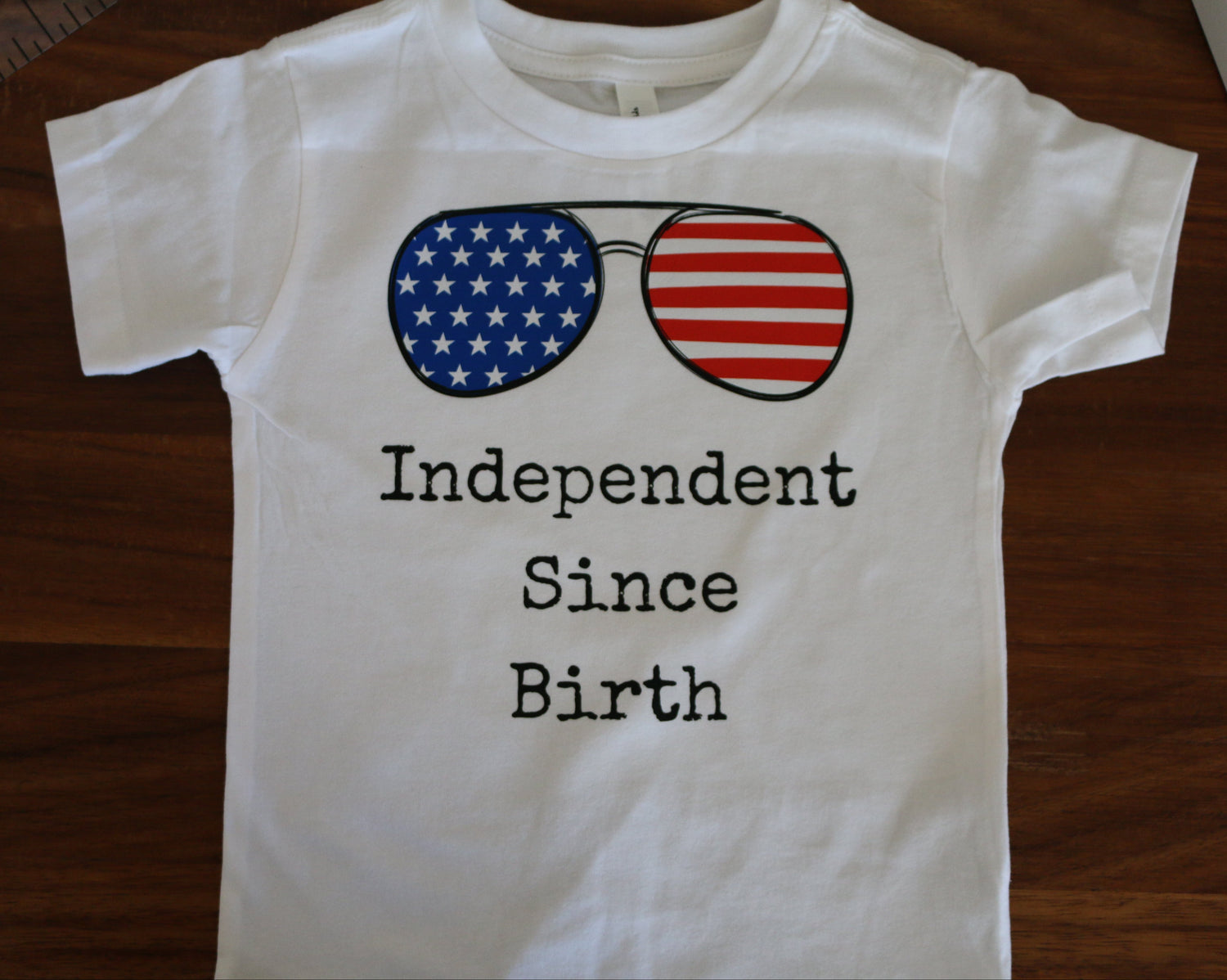 White t-shirt with American flag sunglasses and 'Independent Since Birth' text on a wooden surface