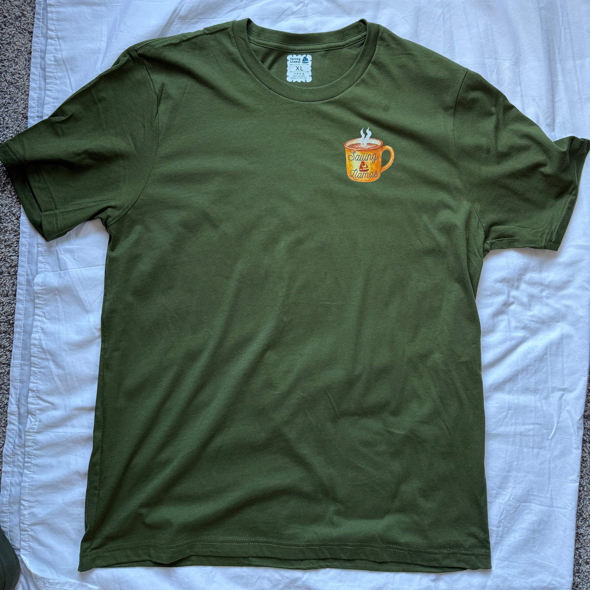 An olive green short-sleeve t-shirt laid flat on a wrinkled white sheet, featuring a small graphic of a yellow coffee mug on the left chest.