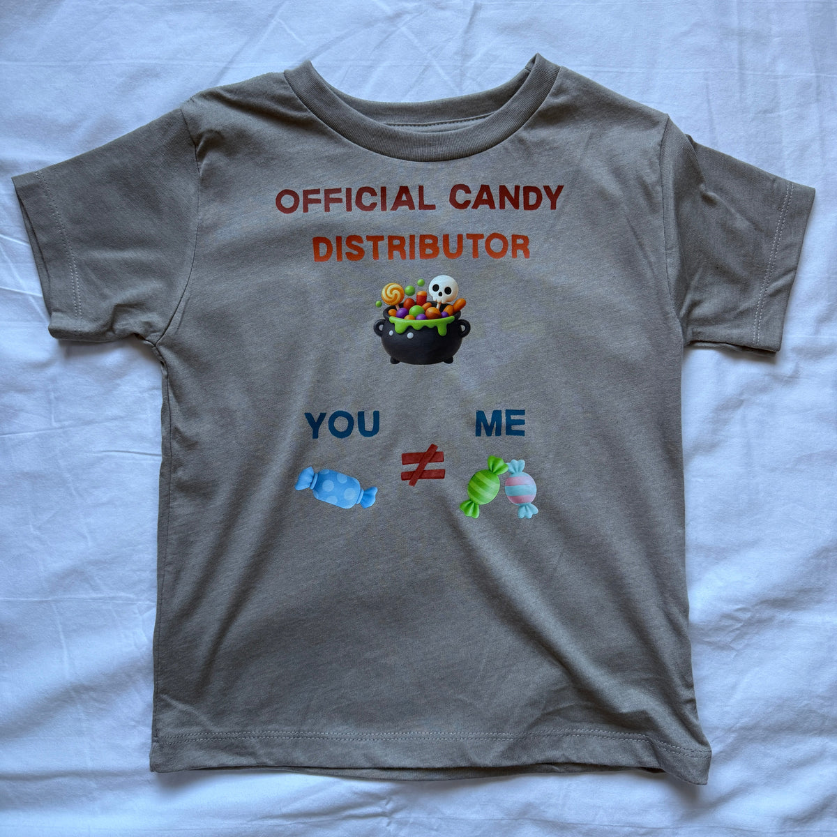 A gray short-sleeve t-shirt laid flat on a white fabric background, featuring a graphic that reads 'OFFICIAL CANDY DISTRIBUTOR' above a pot of candy with characters, and 'YOU != ME' below with candy illustrations.