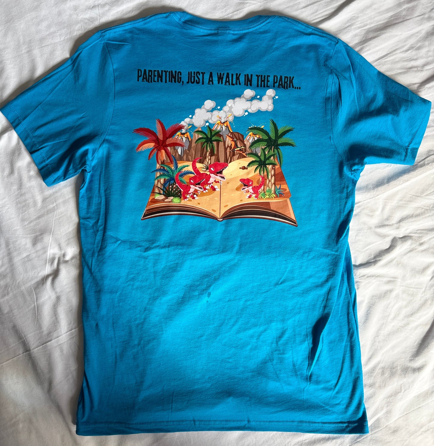 A bright turquoise blue short-sleeve t-shirt laid flat on a wrinkled white sheet. The front of the shirt features a colorful graphic of an open book showing a tropical beach scene with palm trees, a red sun, and clouds, alongside the black text 'PENTINAS, AS A WALK IN THE PARK.