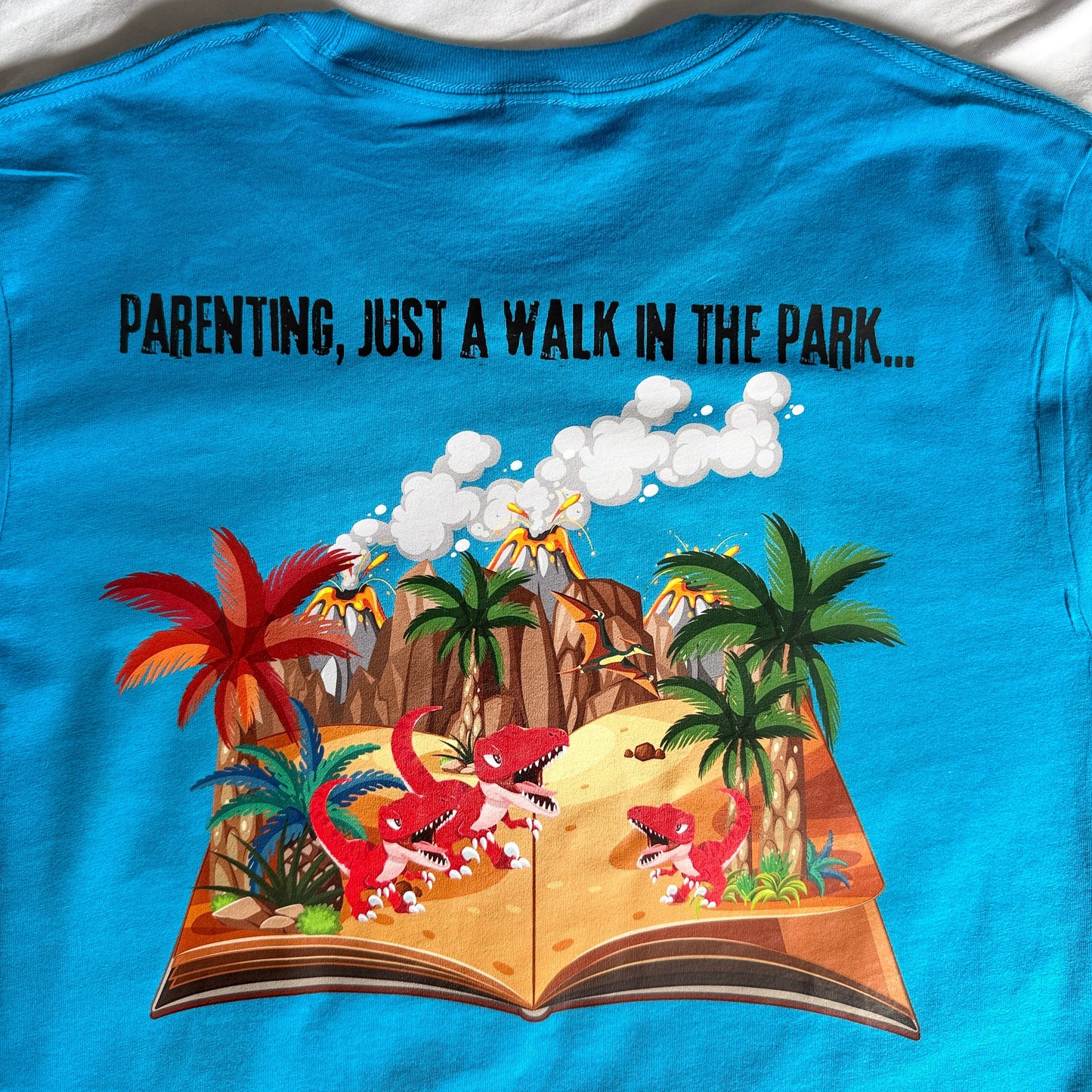 A bright blue graphic T-shirt laid flat on a white fabric background. The shirt features a colorful cartoon illustration of a tropical beach scene with palm trees, red flamingos, and a book opening, alongside black text that reads 'PARENTING, JUST A WALK IN THE PARK.