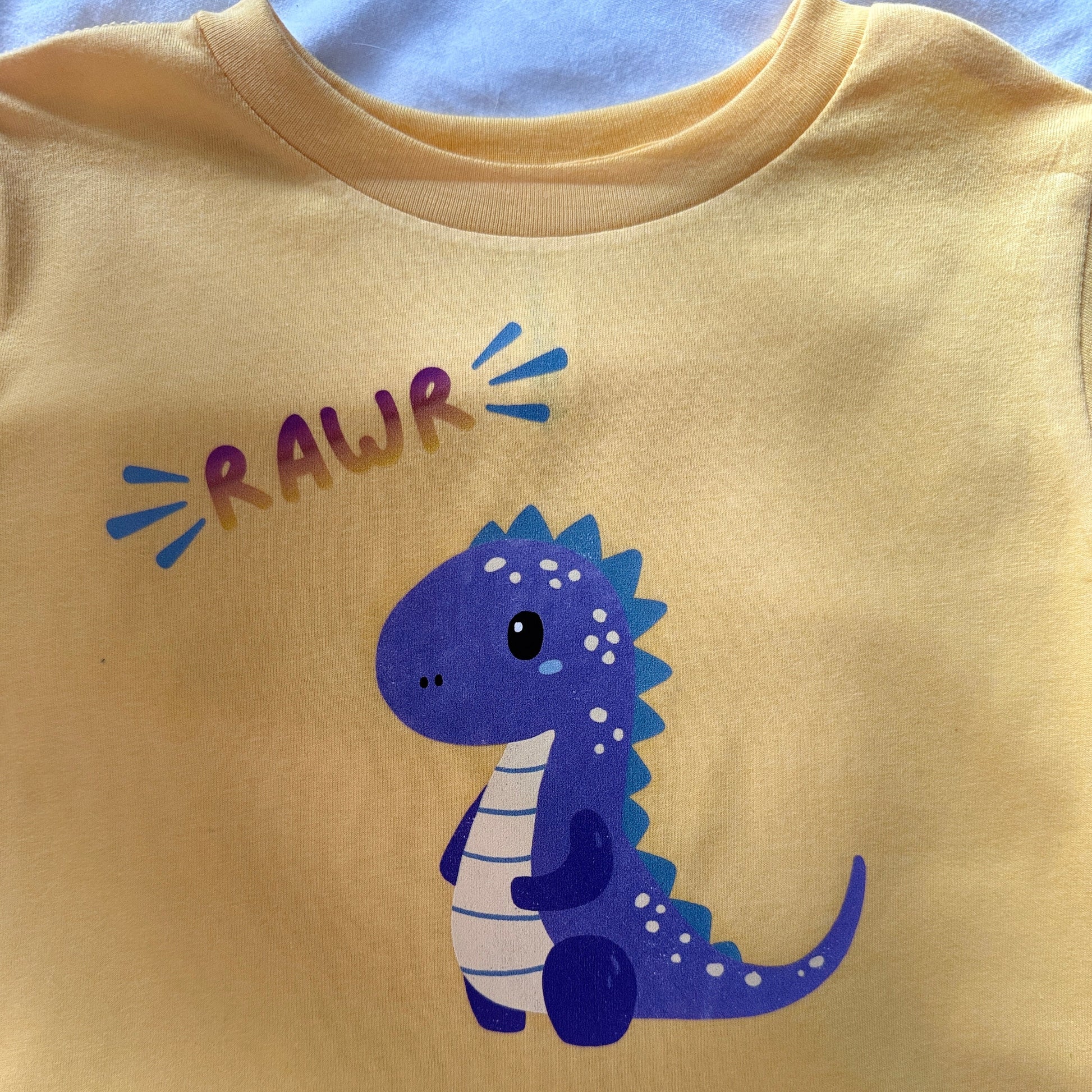 A close-up of a pale yellow toddler t-shirt featuring a cute purple and white cartoon dinosaur with the word 'RAWR' printed above it in purple and blue.