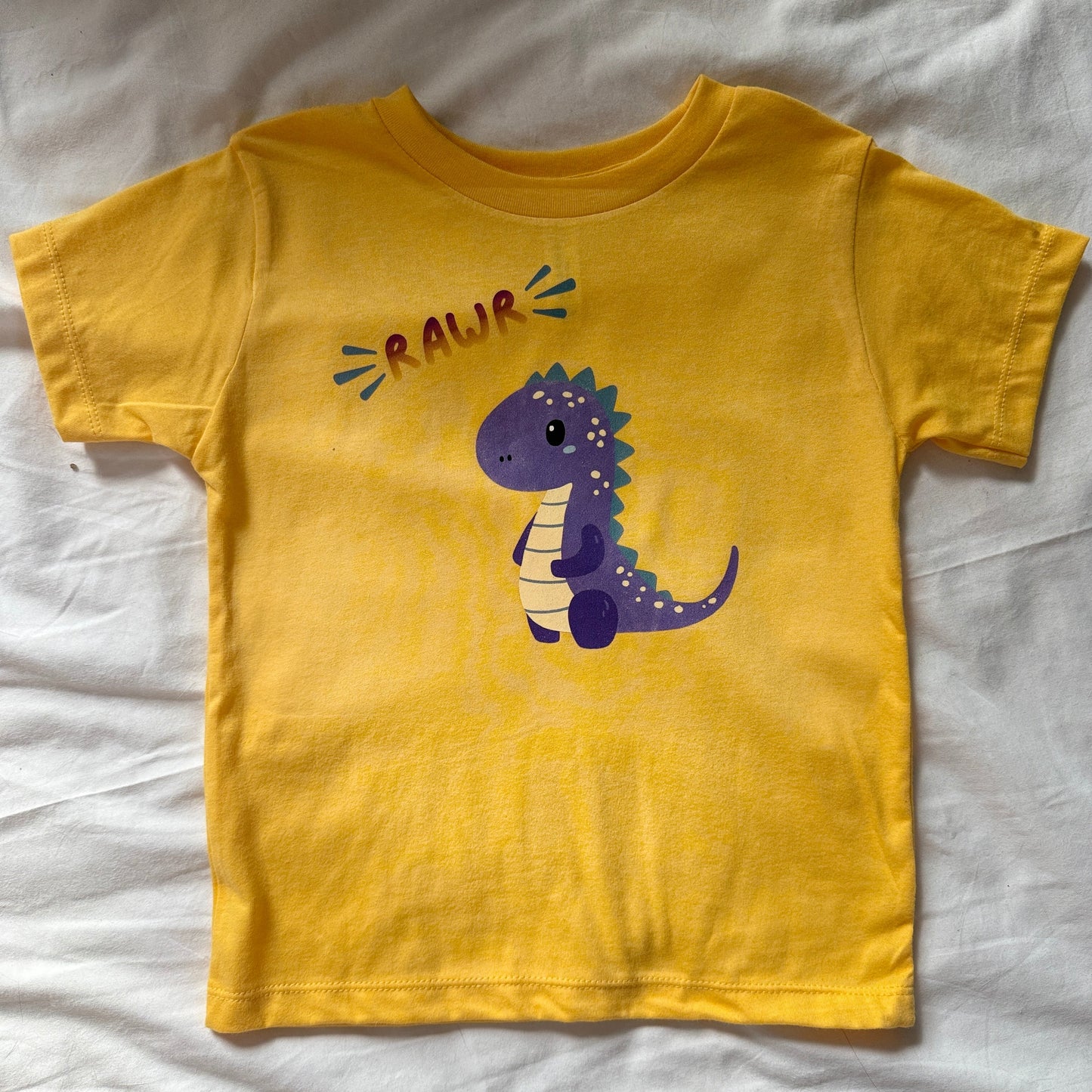 A yellow short-sleeve toddler t-shirt laid flat on a white fabric background, featuring a purple cartoon dinosaur and the word 'RAUR' printed on the front.