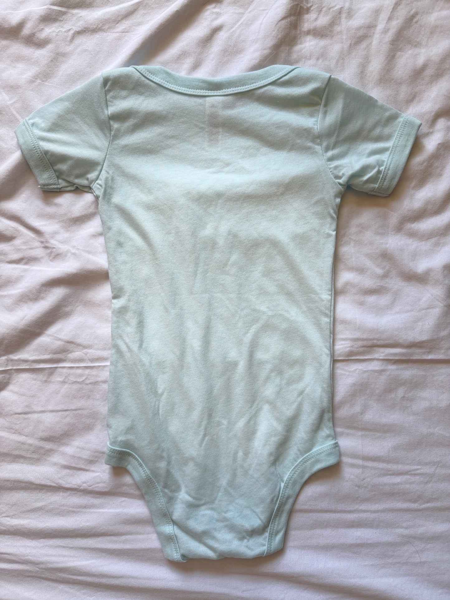A ice blue short-sleeve baby bodysuit laid flat on a wrinkled white sheet.