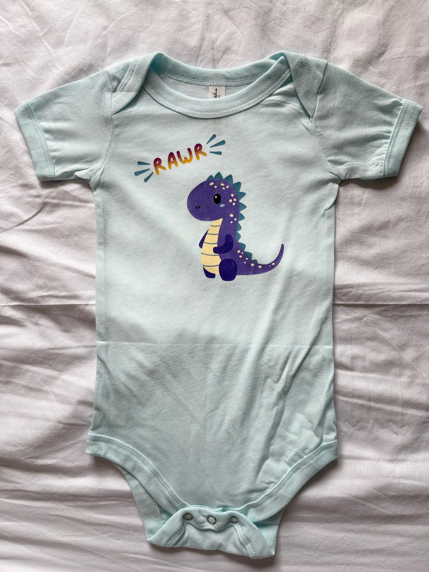 A ice blue baby bodysuit with a purple cartoon dinosaur and the word 'RAWR' printed on the chest, laid flat on a white fabric background.
