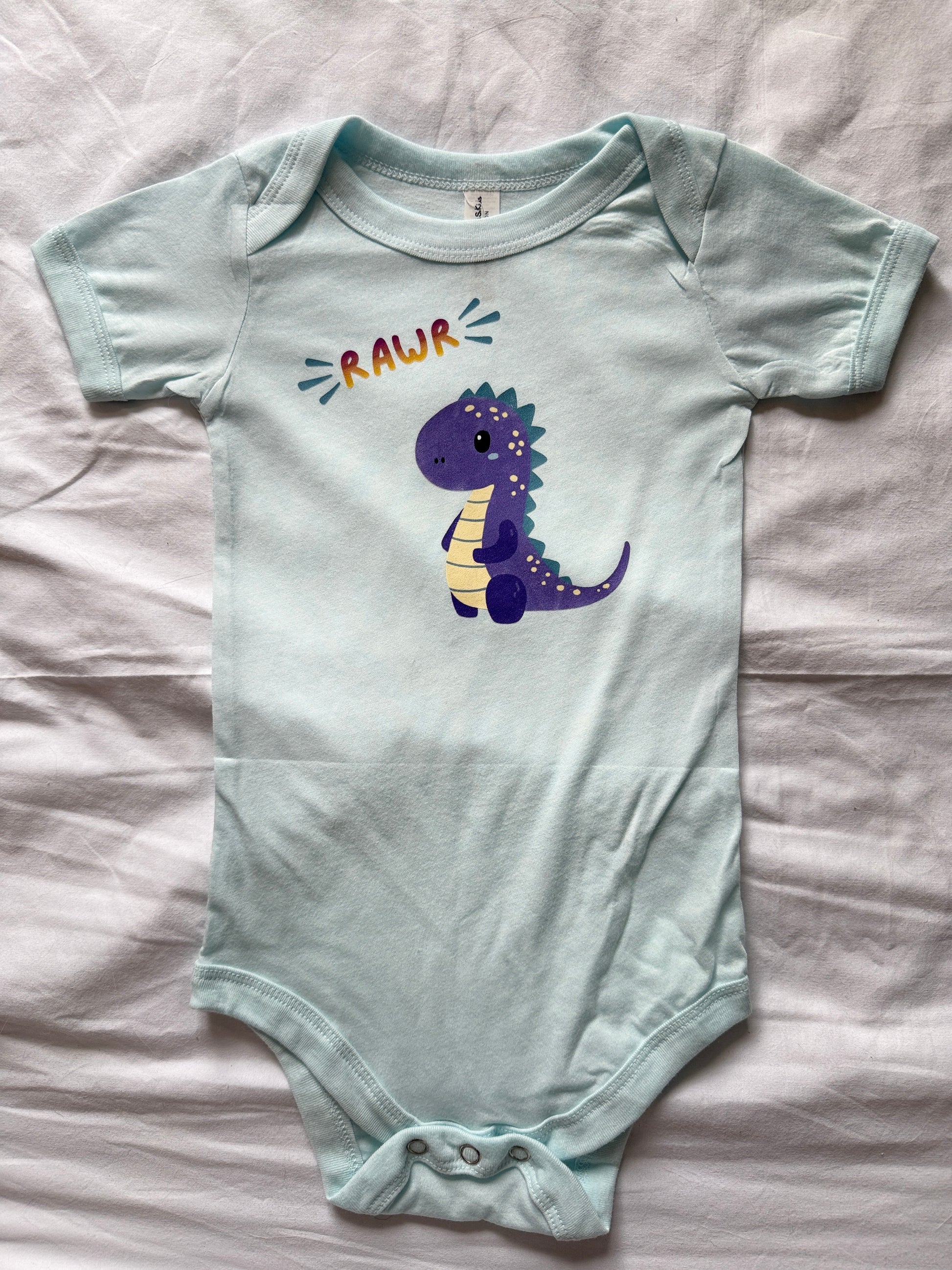 A ice blue baby bodysuit with a purple cartoon dinosaur and the word 'RAWR' printed on the chest, laid flat on a white fabric background.