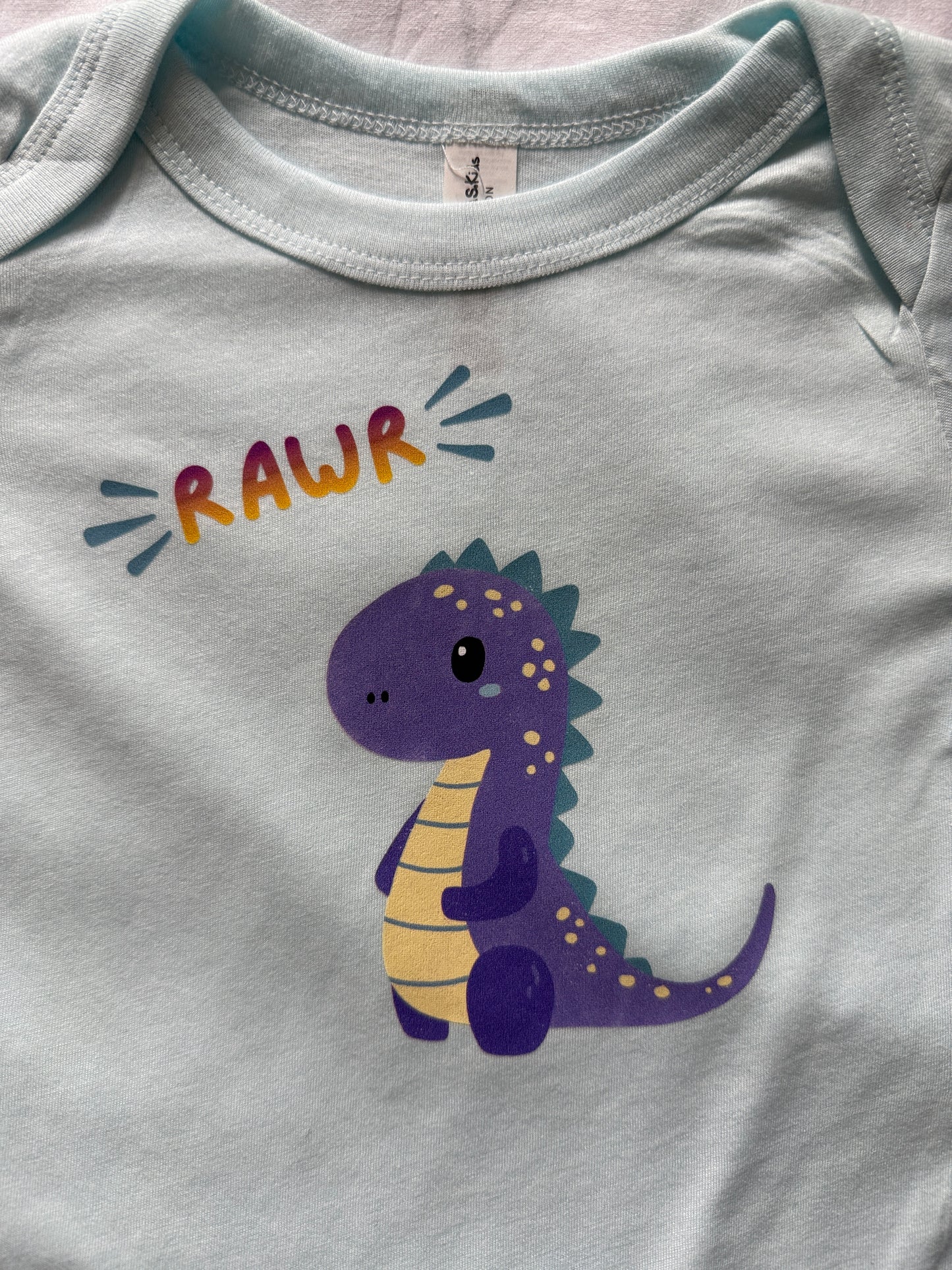 A close-up of a baby bodysuit in a pale icy blue color, featuring a cute purple and yellow dinosaur graphic with the word 'RAWR' printed above it.