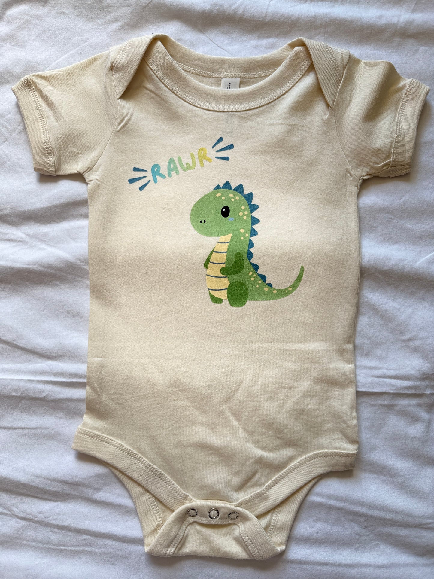 A cream-colored baby bodysuit with a cartoon green dinosaur and the word 'RAWR' printed on the chest, laid flat on a white fabric background.