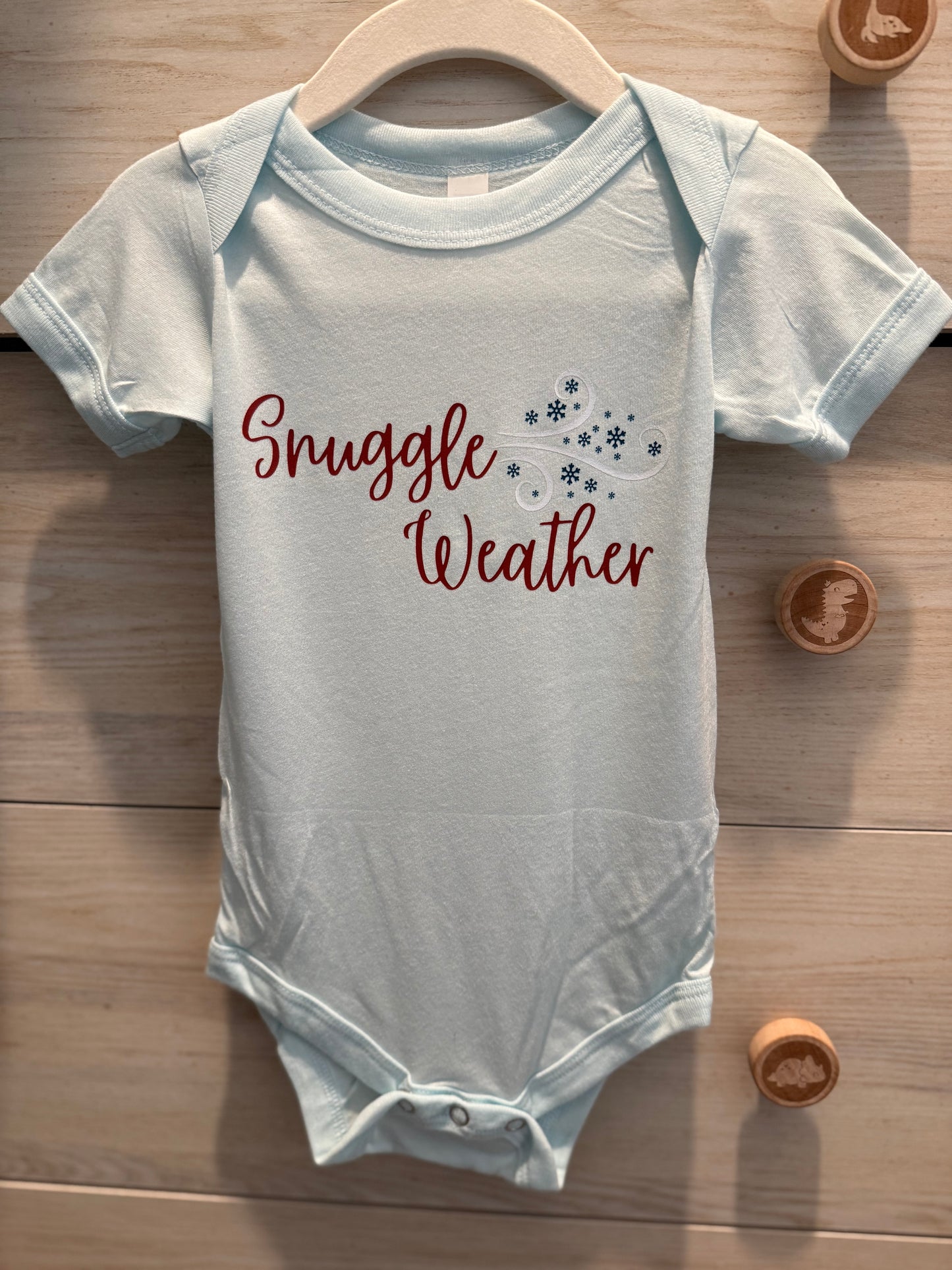 Gray baby onesie with 'Snuggle Weather' text on a wooden surface