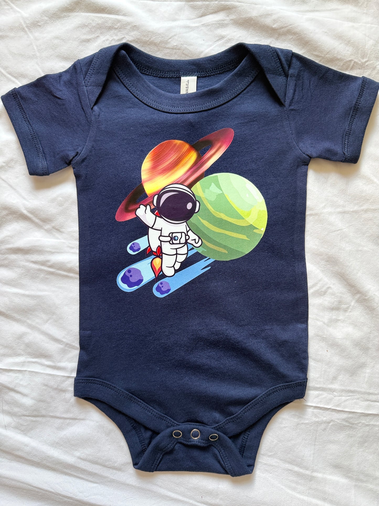 A navy blue baby bodysuit laid flat on a white fabric background, featuring a colorful graphic of an astronaut skiing in front of planets.
