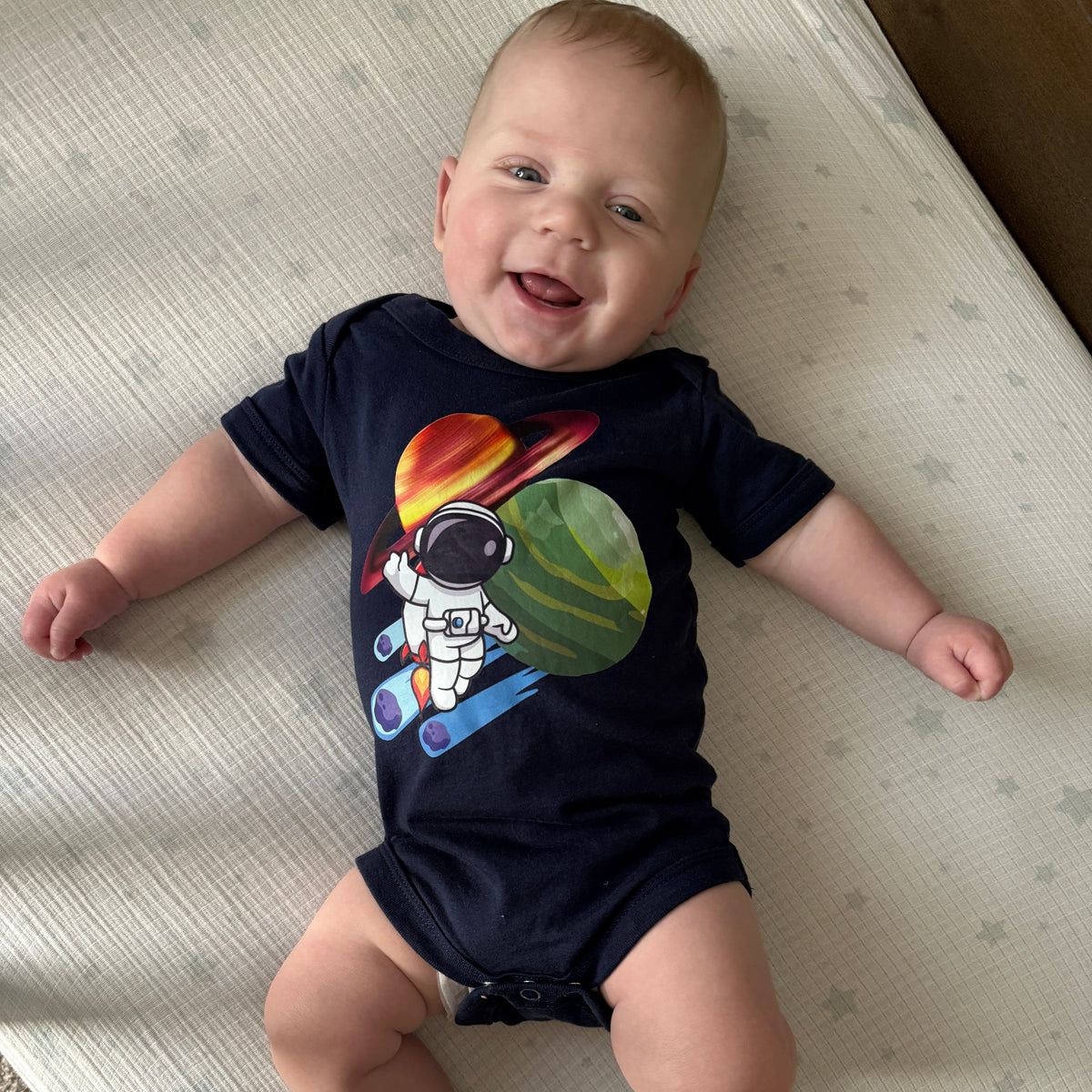 A baby lying on a cream-colored mattress, wearing a navy blue short-sleeve bodysuit with a colorful astronaut graphic on the front.