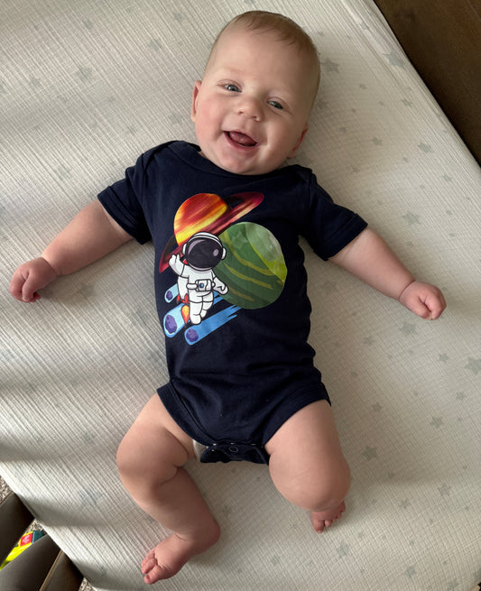 A baby lying on a cream-colored mattress, wearing a navy blue short-sleeve bodysuit with a colorful astronaut graphic on the front.