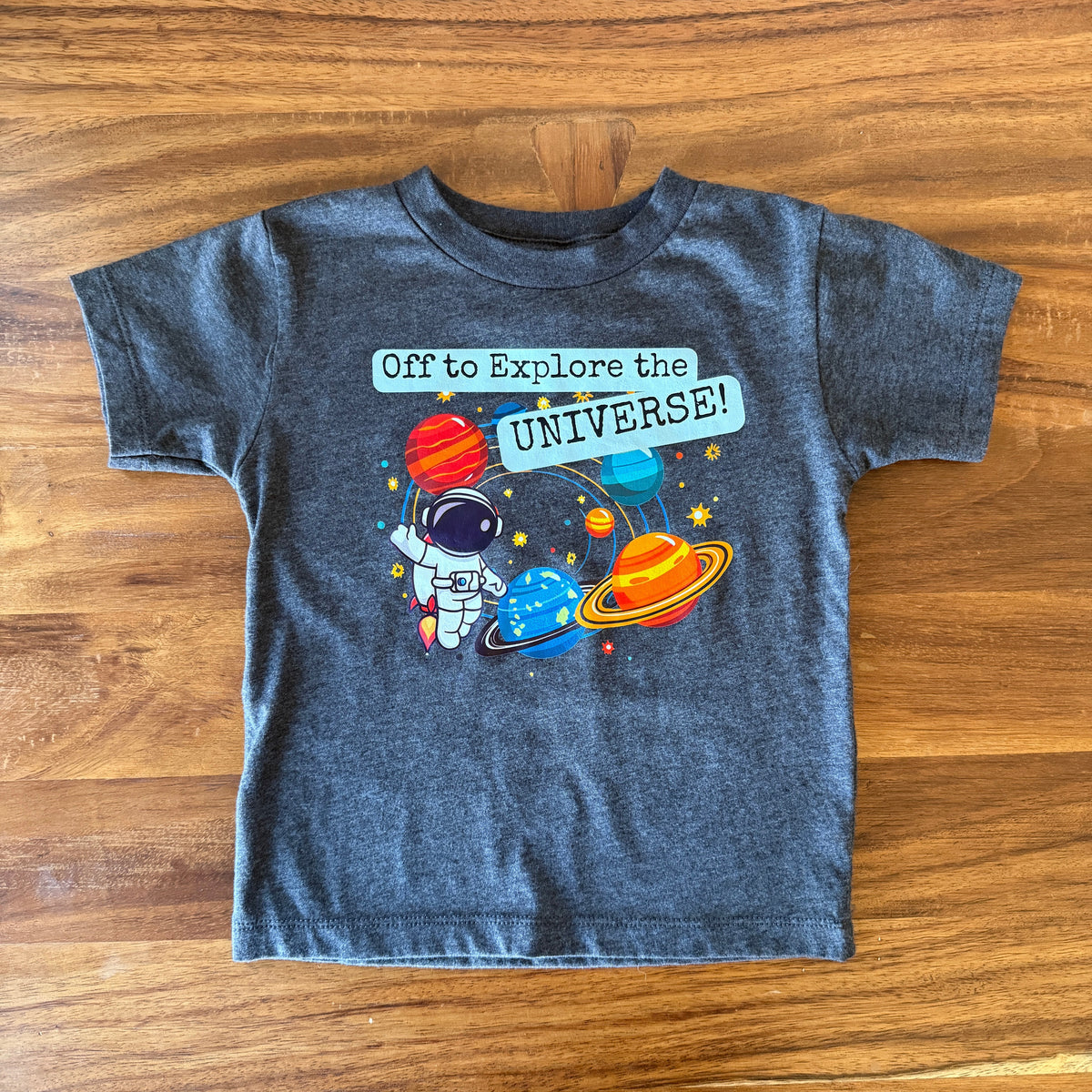 A toddler's dark gray heather t-shirt with a colorful space graphic and the text 'Off to Explore the UNIVERSE!' laid flat on a wooden table.