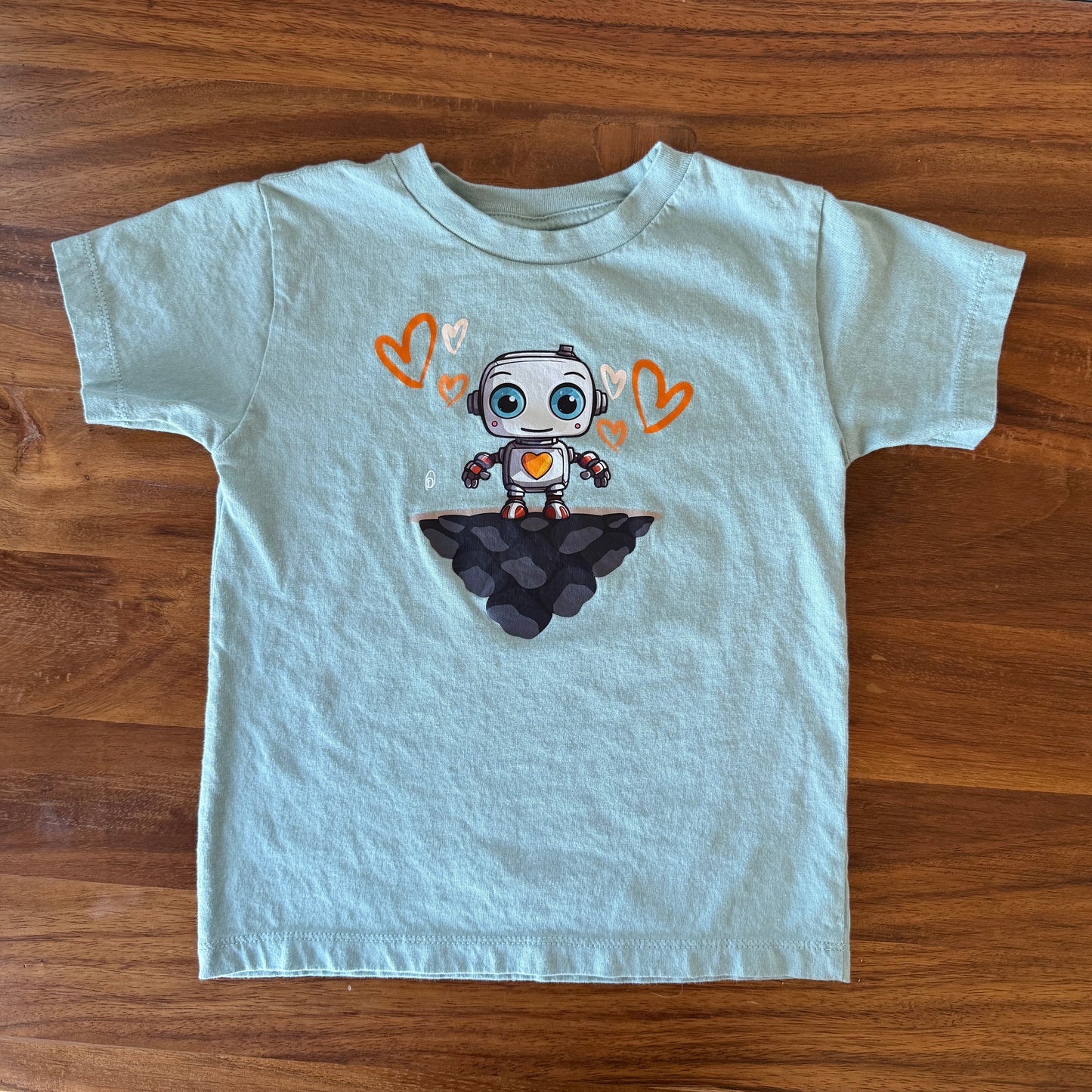 A light blue short-sleeve kids' t-shirt laid flat on a wooden table, featuring a cartoon robot graphic on the chest surrounded by orange hearts.